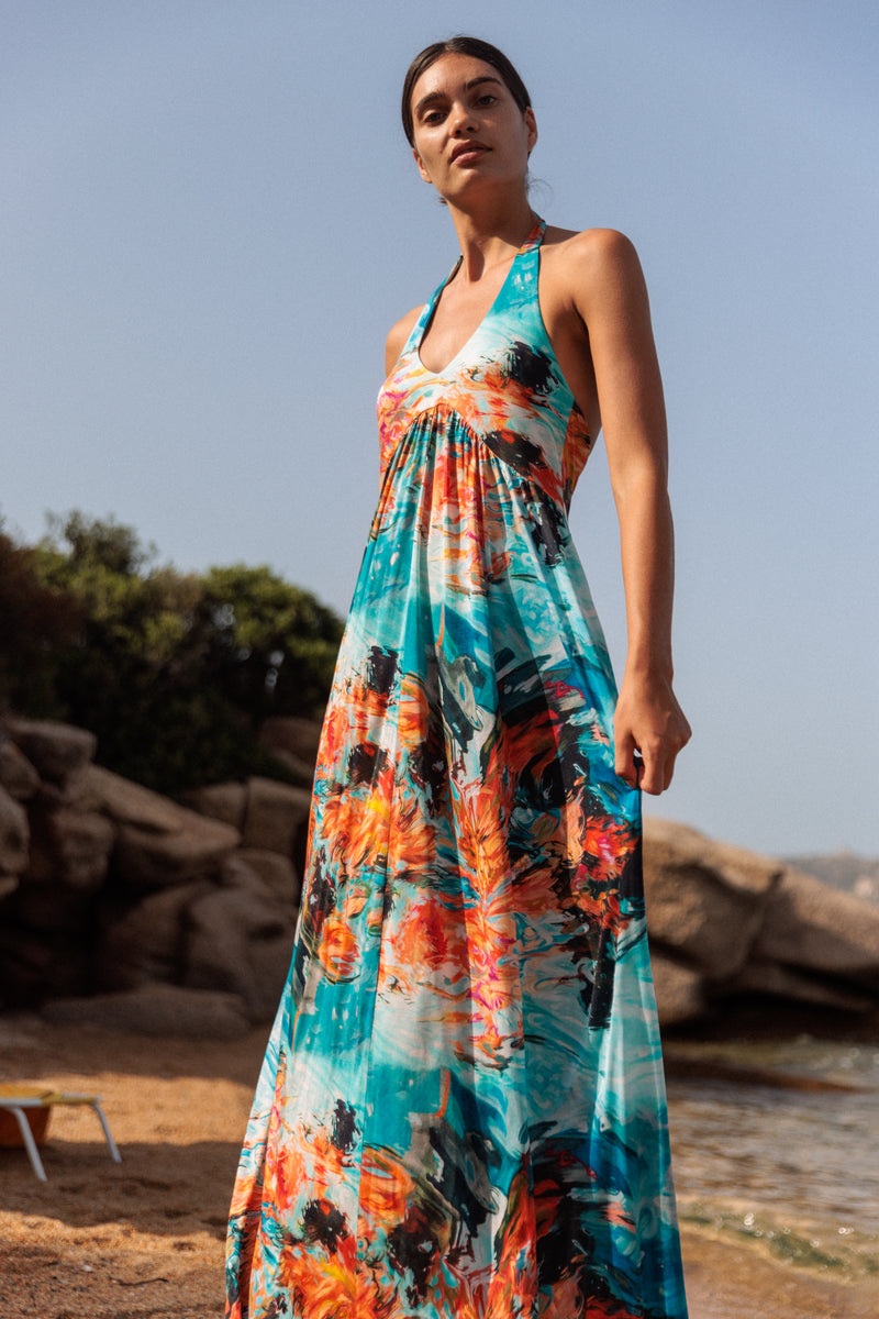 A woman is wearing a Catherine Gee Anna Halter - 7 Girls Dress featuring a vibrant, multicolored abstract pattern with shades of blue, orange, and black. The dress has a halter neckline, and fitted bodice, and flows into a full skirt. The background shows an outdoor setting with rocks, greenery, and a glimpse of water, suggesting a beach or coastal location.