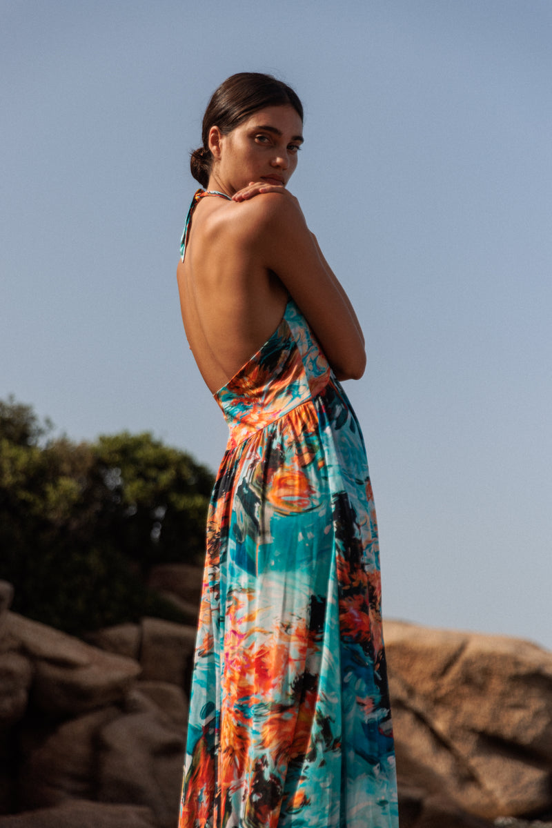 A woman wearing a colorful, backless halter dress with a floral pattern of Catherine Gee Anna Halter Dress - 7 Girls. The dress is long and flowing, featuring a mix of vibrant colors including blue, orange, and black. The person is standing outdoors, with a background of rocks and greenery under a clear sky. The image showcases the elegant design and vibrant colors of the dress, making it visually appealing and relevant for fashion enthusiasts.