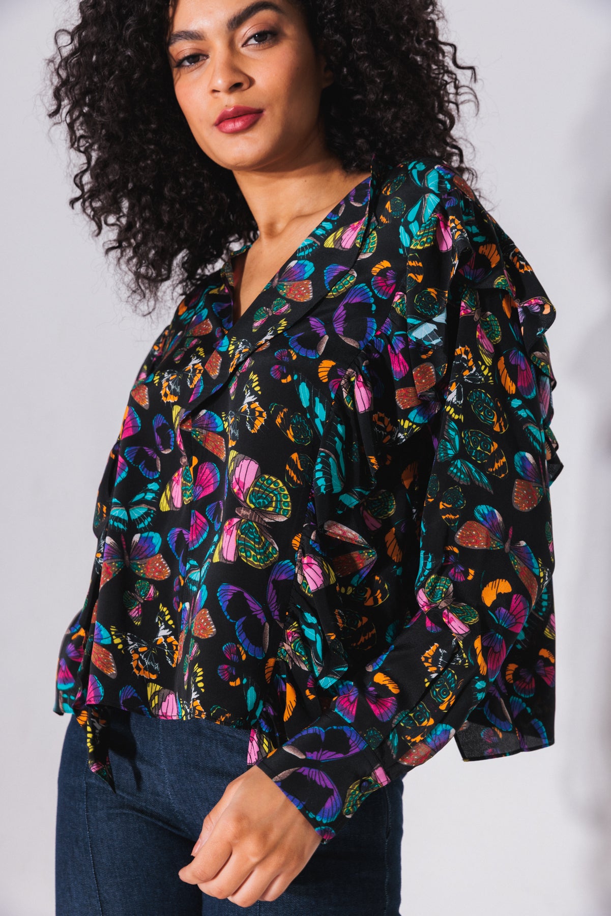 A woman is wearing the Catherine Gee Bella Blouse - The Tracy features a vibrant, multicolored butterfly print on a black background, with a loose fit, long sleeves, and a V-neckline paired with dark blue jeans. The image focuses on the upper body, highlighting the blouse's design and pattern.