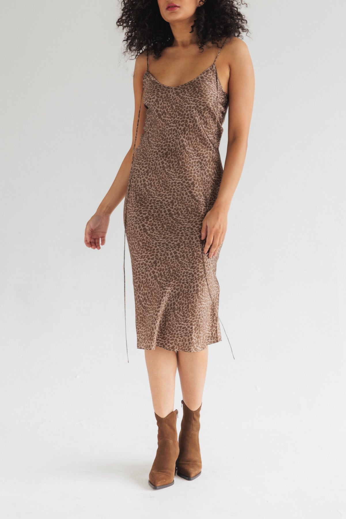 A woman wearing the Catherine Gee Emma Elegant Leopard Slip Dress paired with brown ankle boots adds a stylish touch. This chic, mid-length dress features a bold leopard print pattern and thin spaghetti straps, and has a relaxed fit, falling gracefully to mid-calf. The silky fabric gives it a luxurious and elegant feel, making it suitable for both casual and semi-formal occasions.