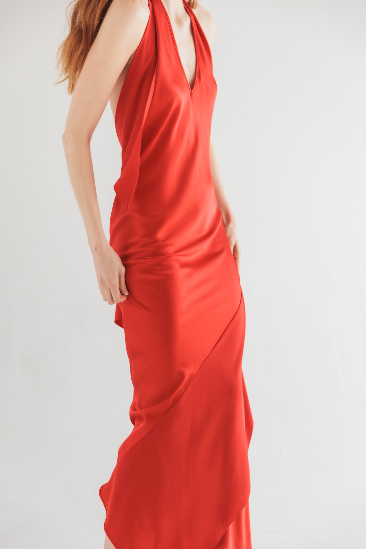 A woman wearing the Catherine Gee Diane High-Risk vibrant red dress features a sleeveless design with a deep V-neckline. The smooth, silky fabric drapes elegantly over the body, creating a fitted silhouette that flares slightly at the bottom. She poses with hands resting on the sides of the dress, highlighting its sleek and flowing design, making it a sophisticated and stylish choice for formal occasions.