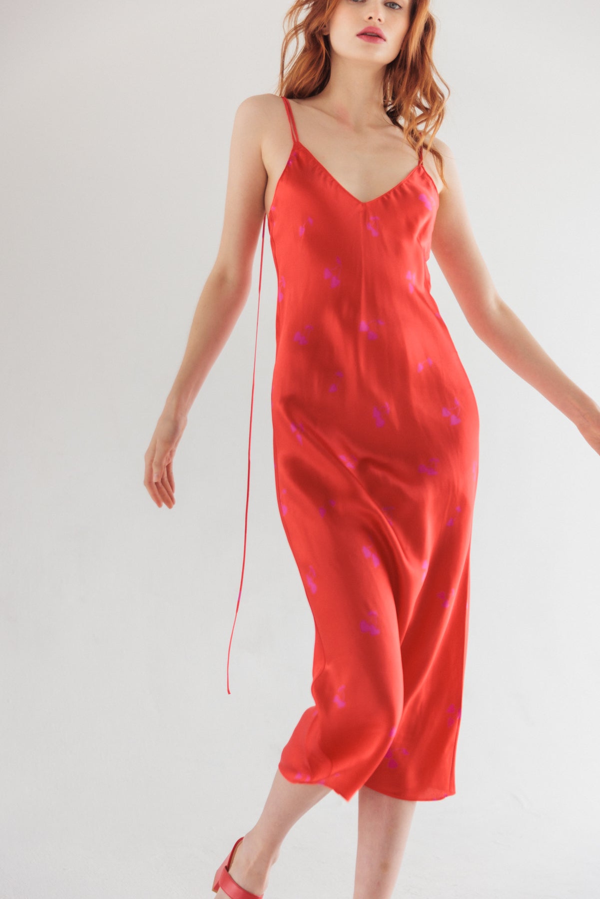 A woman posing gracefully wearing a Catherine Gee Emma Cherry Heart Slip Dress. The dress features a vibrant red color with a pattern of delicate purple hearts scattered throughout. It has a V-neckline and thin spaghetti straps, creating an elegant and timeless look. The fabric appears silky and smooth, draping beautifully over the body highlighting the dress's fluid silhouette and sophisticated design.