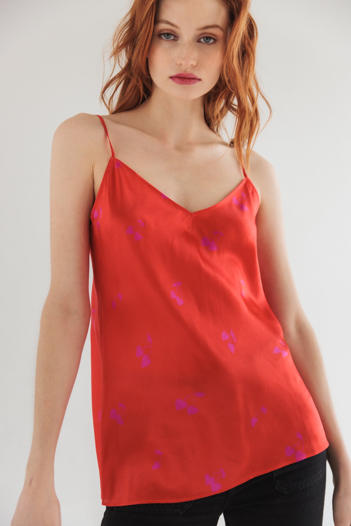 A woman wearing a red Catherine Gee Basic Cami - Cherry Heart with thin straps and a V-neckline, featuring a subtle pattern of small purple hearts paired with dark pants. The light-colored, plain background makes the red camisole stand out, focusing on the upper body and showcasing the design and fit of the camisole.