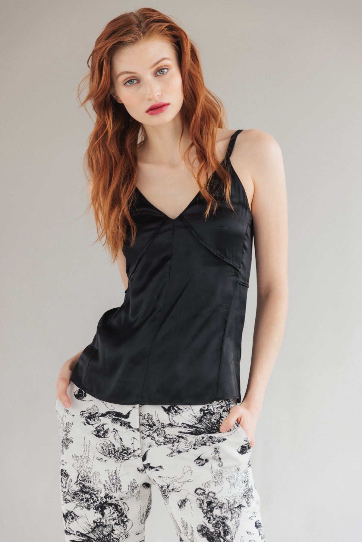 A woman wearing a Catherine Gee Catherine black Tank top with thin straps and a V-neckline paired with white pants featuring a black floral pattern. The person has long, wavy red hair and is posing with hands in the pockets of the pants against a plain background.