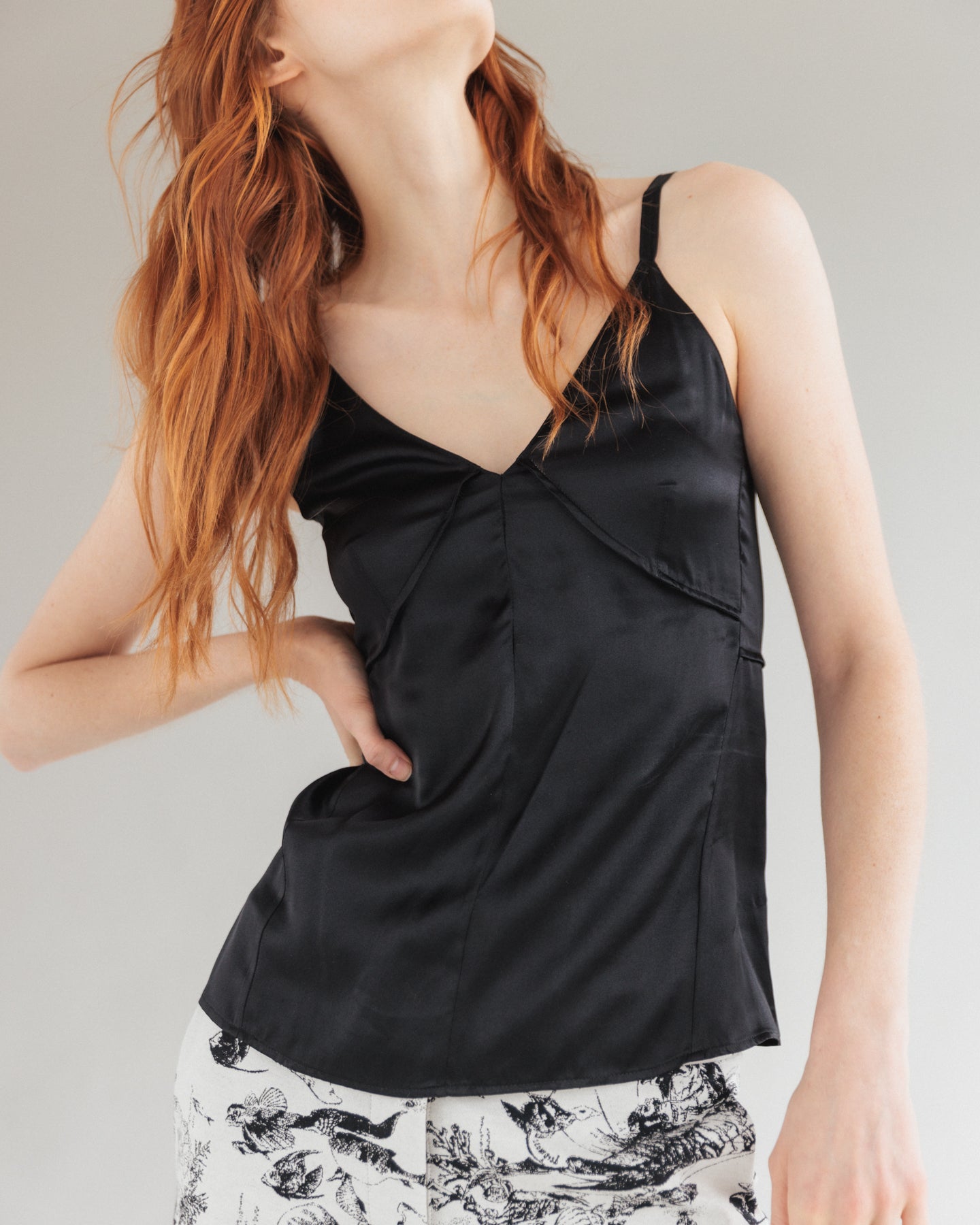 A woman with long red hair is wearing a black Catherine Tank satin camisole top with thin straps and a V-neckline by Catherine Gee. The top has a fitted design with seam details. The person is also wearing white shorts with a black intricate pattern. The background is plain and neutral, highlighting the outfit.