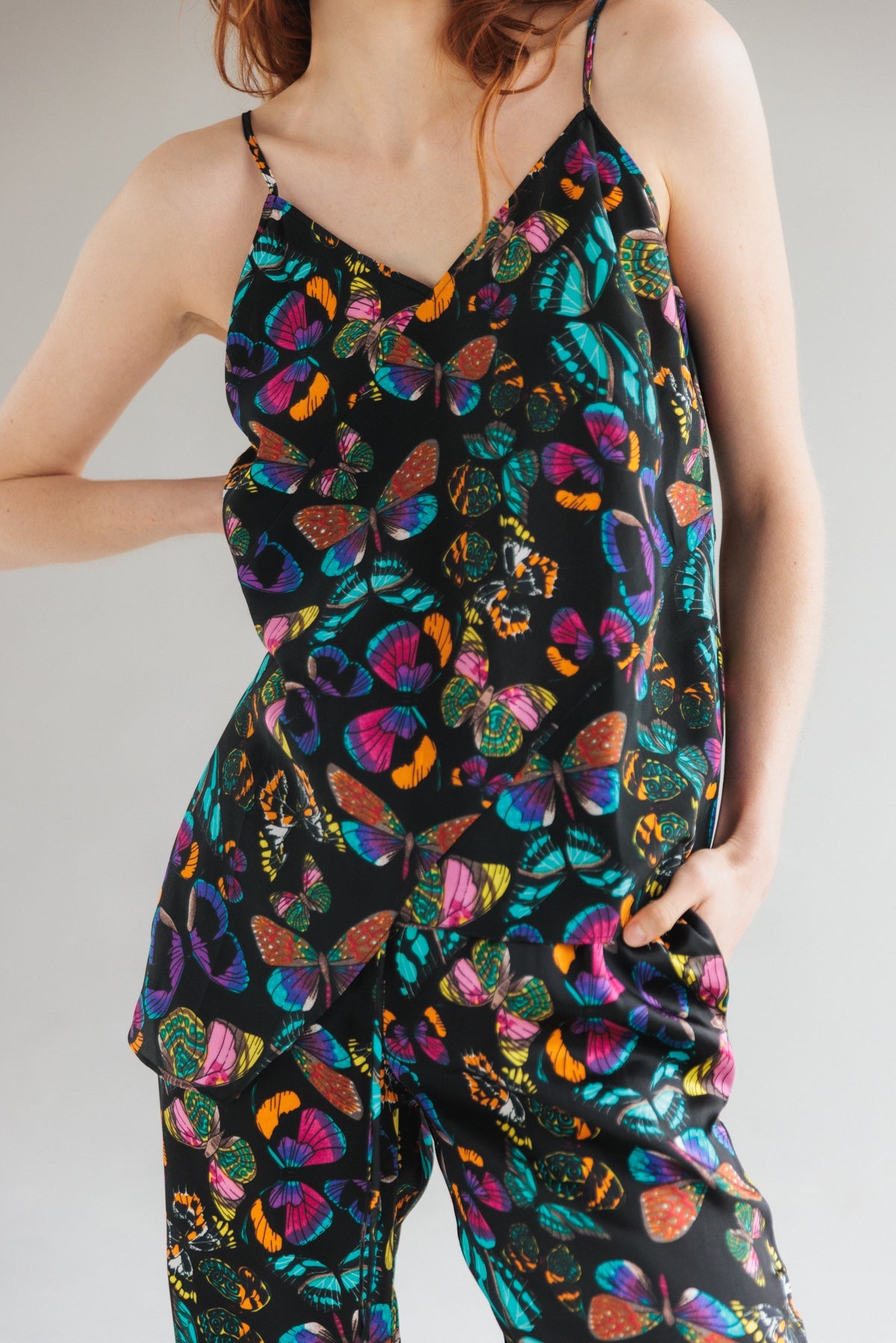 A woman posing with one hand on her hip and the other arm relaxed by her side is wearing a Catherine Gee Alexa Tank in the 'The Tracy' design. The tank top features a vibrant butterfly print with various colors including blue, purple, orange, and green on a black background, with thin straps and a V-neckline. 