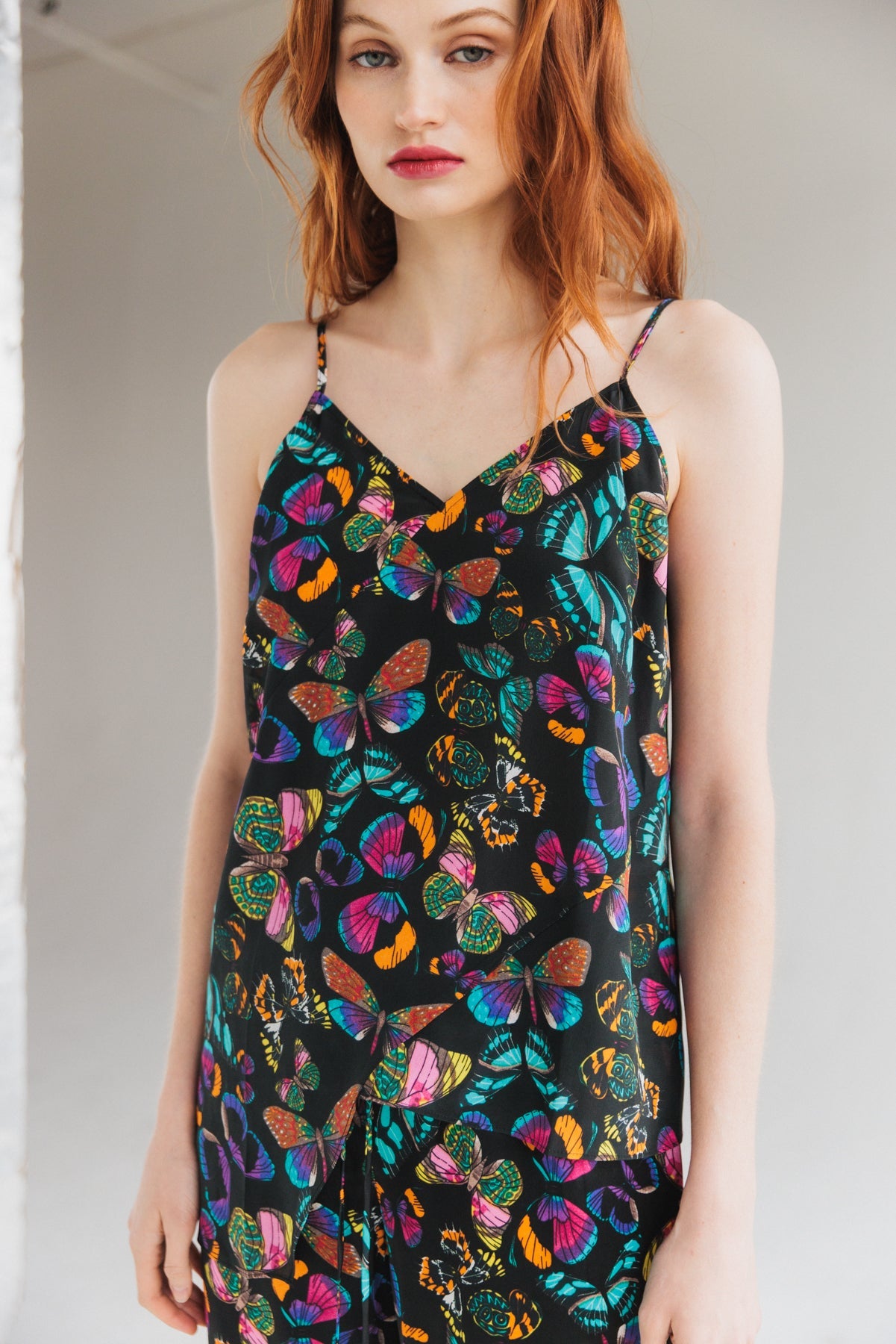 Woman wearing a Catherine Gee Alexa Tank - The Tracy in the neutral and minimalistic background, featuring a black background with a vibrant butterfly print in various colors including blue, purple, orange, and green. The tank top has thin straps and a V-neckline.