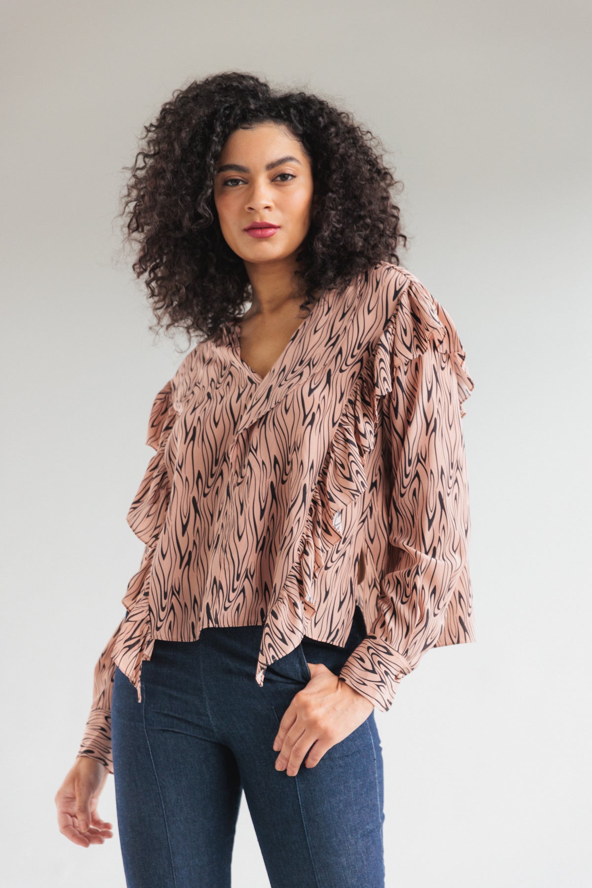A woman is wearing a Catherine Gee Bella Blouse - Wishbone features a light pink base with a black abstract wishbone-like design. It has a V-neckline, long sleeves with ruffled details, and a loose fit paired with dark blue jeans. The background is plain and light-colored, emphasizing the blouse's unique and stylish design.