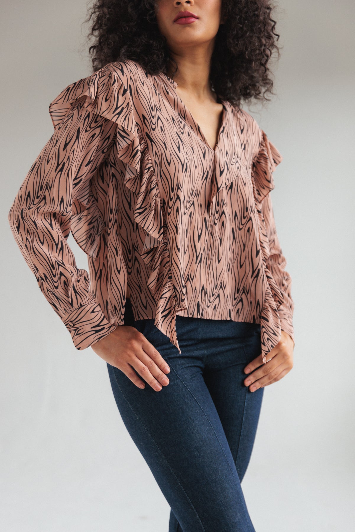 A woman wearing a Catherine Gee Bella Blouse - Wishbone. The blouse features a pink base with an abstract black pattern, long sleeves, and ruffled details along the front and shoulders. She is also wearing dark blue jeans in a plain, light color background that highlights the blouse's unique design.