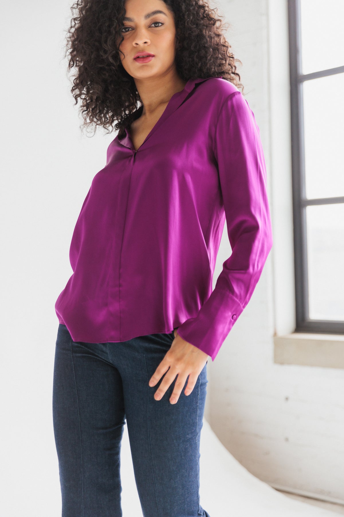 A woman wearing a Catherine Gee Daria French Cuff Silk Blouse Electric Orchid. The blouse has a V-neckline and long sleeves with French cuffs paired with dark blue jeans. The background features a large window with natural light.