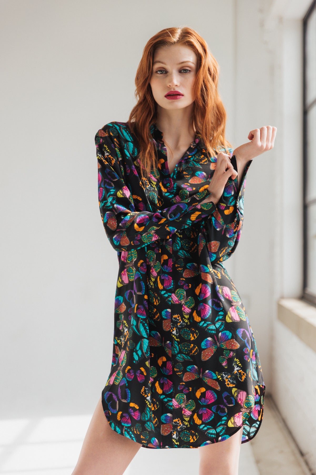 A woman wearing the Catherine Gee Daria Silk Shirt The Tracy dress features a vibrant, multicolored butterfly with various shapes and designs on a dark background. It has long sleeves, a loose, comfortable fit, and falls to mid-thigh length. She's standing in a well-lit room with a large window, which adds a natural light effect to the scene.