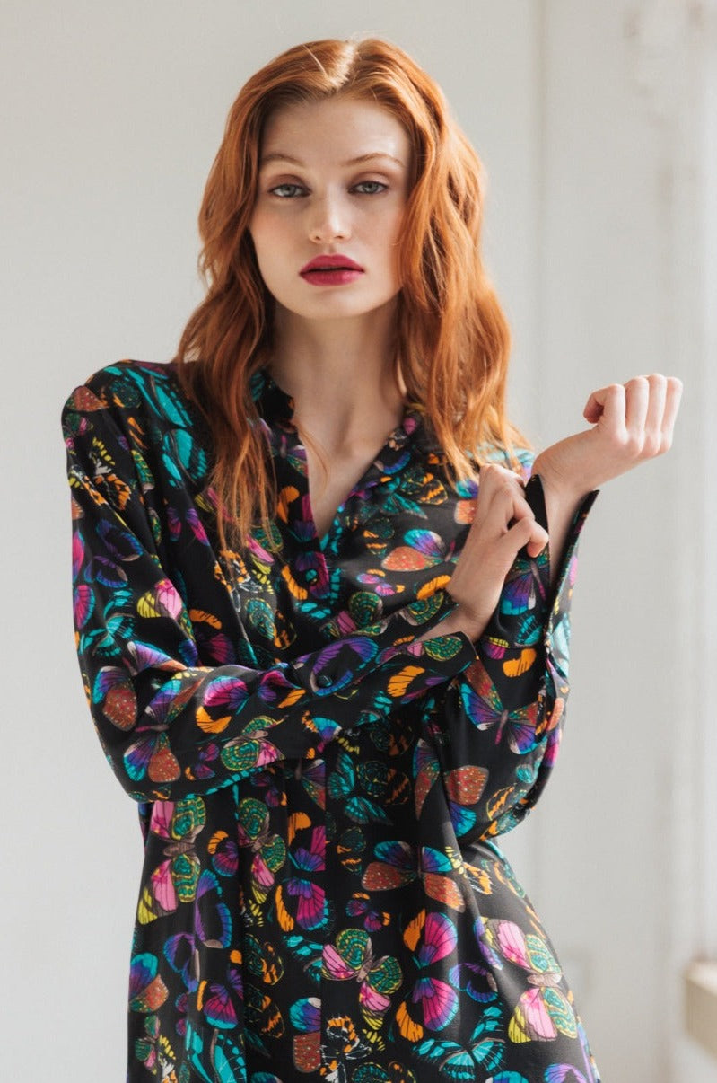 A woman posing with one hand near the face and the other hand adjusting the sleeve, wearing the Catherine Gee Daria French Cuff Silk The Tracy blouse features a vibrant and colorful pattern with various shapes and designs, including butterflies and abstract elements. The fabric appears to be silky and smooth, and the blouse has a loose, comfortable fit with long sleeves and French cuffs highlighting the blouse's elegant and stylish design.