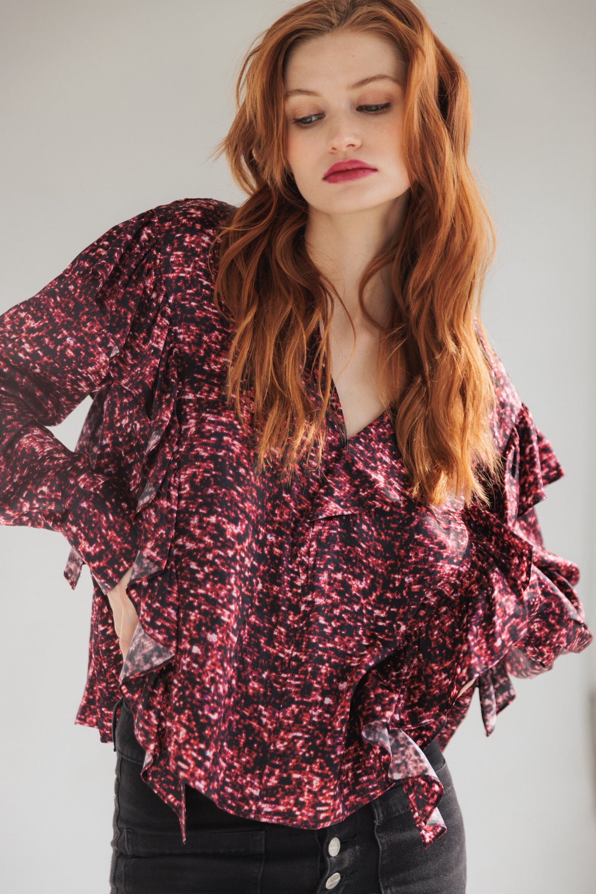 A woman is wearing a Catherine Gee Bella Blouse - Red Sequin features a vibrant red and black speckled pattern with ruffled details along the sleeves and front. She has long, wavy hair and is posing with hands on hips, wearing dark high-waisted pants with visible buttons.