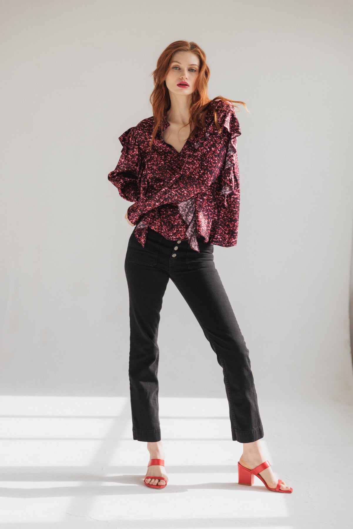 A woman is wearing a Catherine Bella Blouse - Red Sequin standing with one hand on the hip and the other relaxed by the side in a plain, light-colored wall background. The blouse features a vibrant red and black speckled pattern with ruffled details along the sleeves paired with black high-waisted pants and red open-toe heels.
