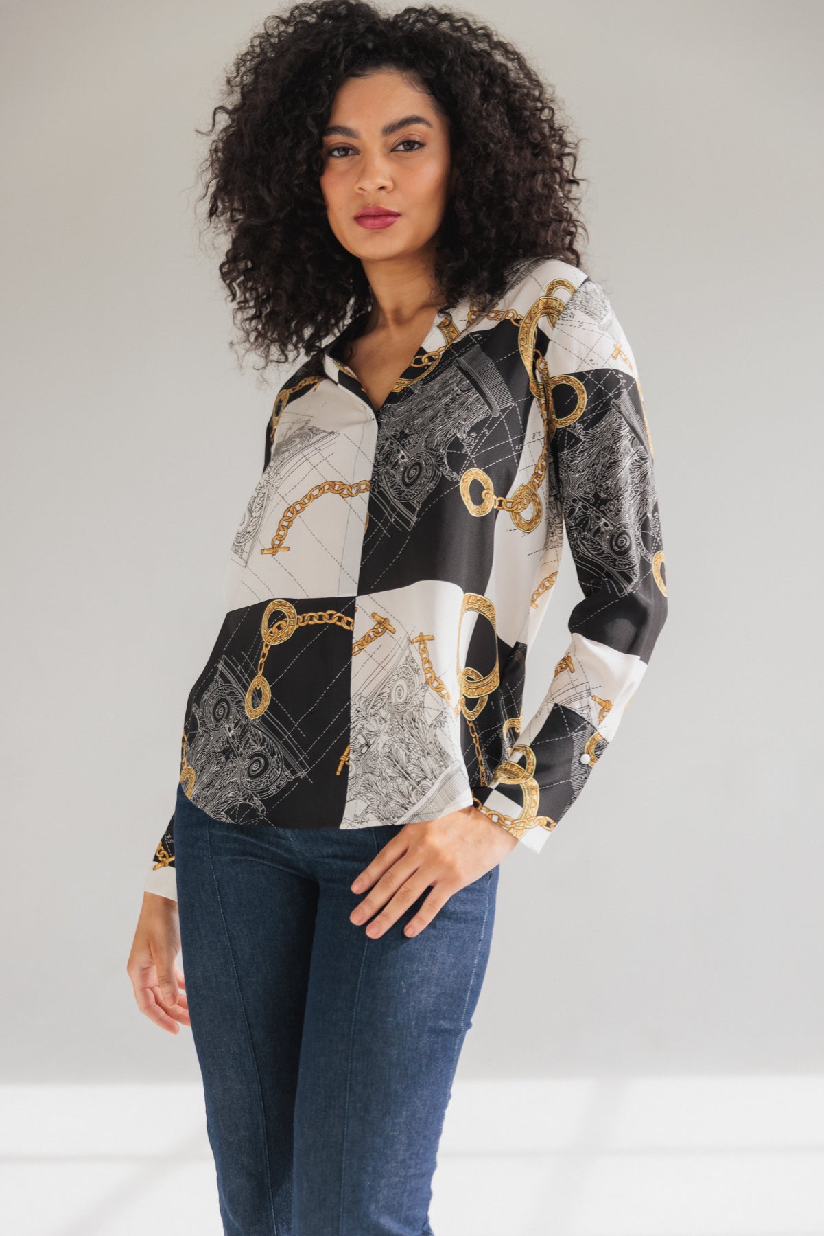 Woman wearing a Catherine Gee Daria French Cuff Silk Blouse Athens, featuring a mix of black, white, and intricate gold chain designs paired with dark blue jeans. The background is plain and light-colored, highlighting the detailed patterns and luxurious feel of the blouse.