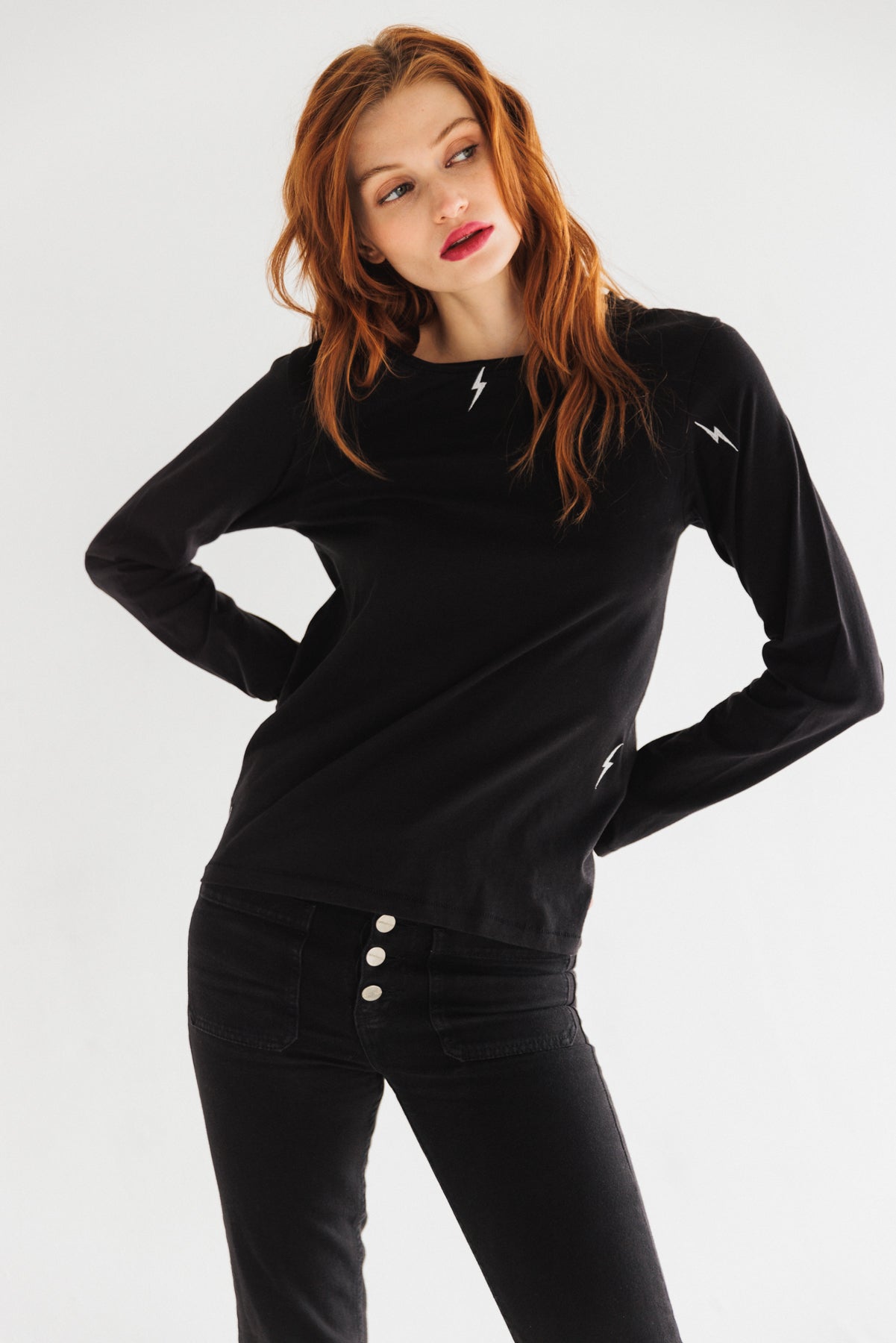 A woman wearing a black Catherine Gee Embroidered Cotton Long Sleeve Bolt t-shirt features small white lightning bolt embroidery on the chest, sleeve, and near the waist paired with black pants that have three visible buttons on the front. The plain white background highlights the minimalist yet stylish design of the t-shirt, emphasizing its subtle embroidered details.