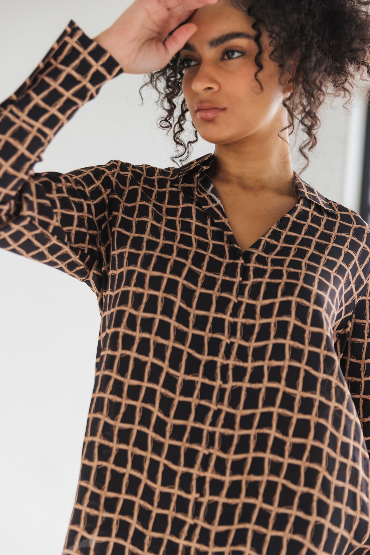 A woman wearing a Catherine Gee Daria French Cuff Silk Recoleta blouse features a black base color with a grid-like pattern of intersecting beige lines. It has a V-neckline, long sleeves with French cuffs, and a smooth, silky texture that highlights its luxurious nature. The image focuses on the upper body, emphasizing the elegant design and intricate pattern of the blouse, perfect for a sophisticated, polished look.