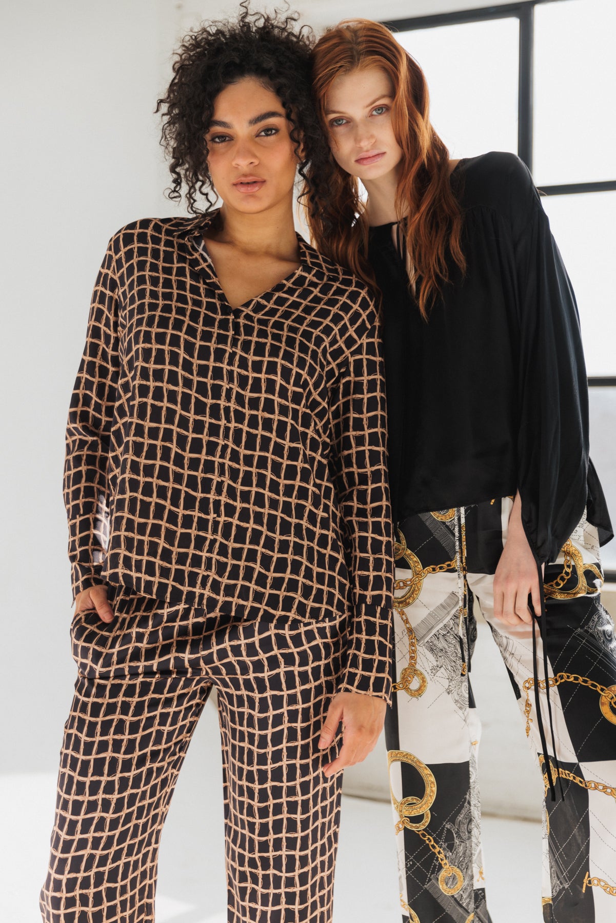 Two women standing side by side. The individual on the left is wearing a matching set of a long-sleeved shirt and pants with a black and beige grid pattern, a Catherine Gee Daria French Cuff Silk Recoleta Blouse, featuring a relaxed fit, classic collar, and French cuffs, showcasing a modern and sophisticated design highlighting the luxurious nature of the material.