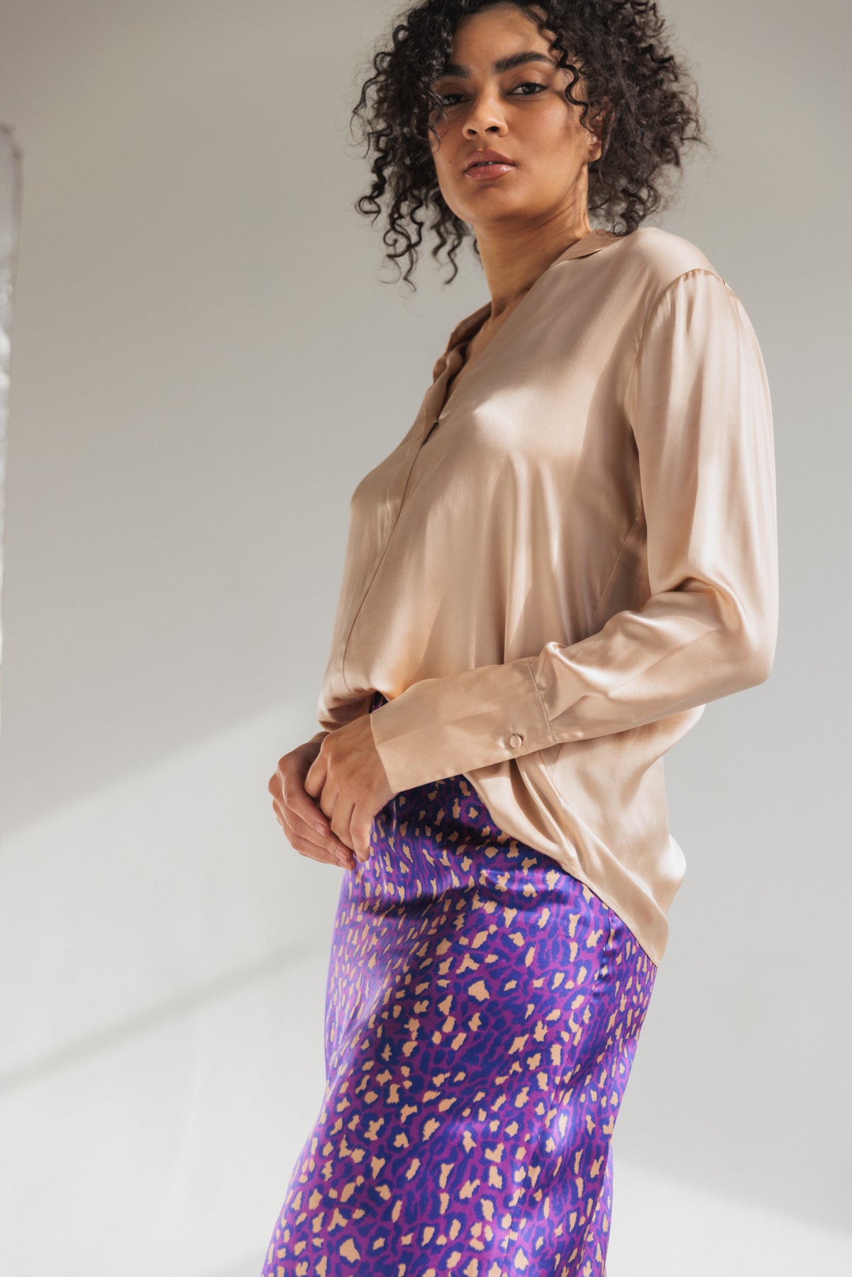 Woman wearing a Catherine Gee Daria French Cuff Silk-Champagne blouse paired with a vibrant purple skirt with a colorful pattern on a plain neutral background.