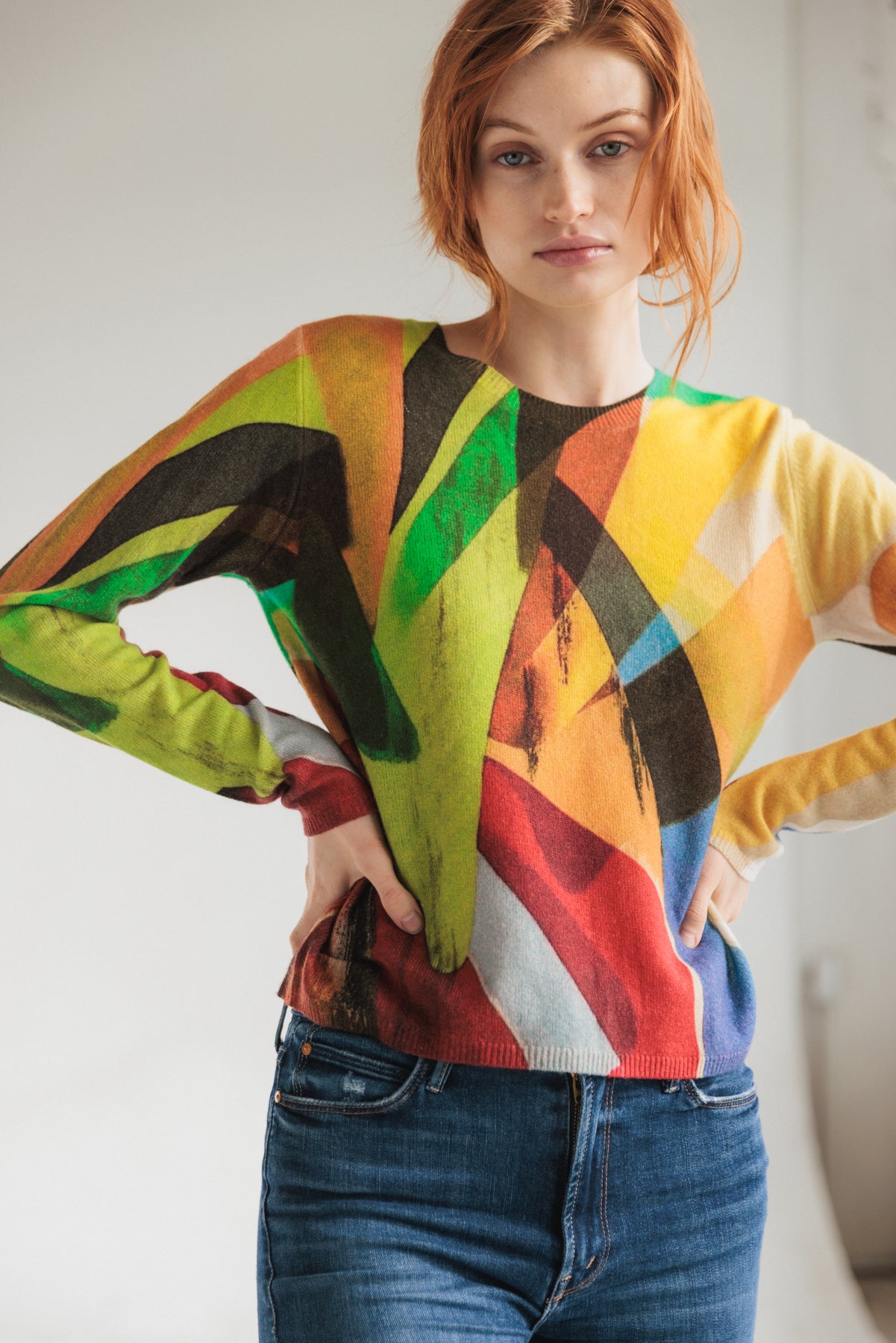 A woman wearing a Catherine Gee Crewneck Cashmere Sweater - Stella Print features a vibrant, abstract design with a mix of colors including green, red, yellow, blue, and black paired with blue jeans and has hands on hips.