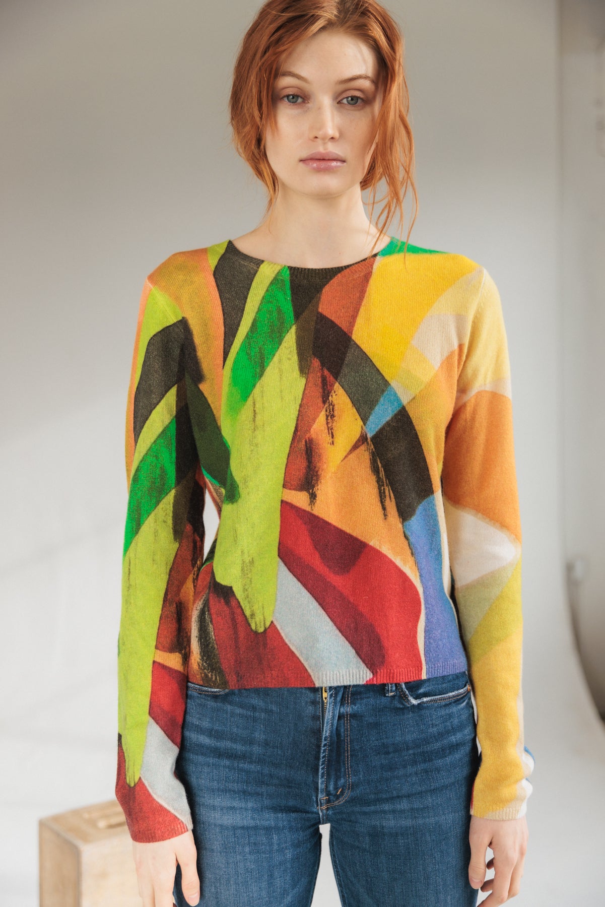 A woman is wearing a Catherine Gee Crewneck Cashmere Sweater - Stella Print design features a vibrant, abstract pattern with bold colors including green, red, yellow, blue, and black paired with blue jeans. The background is neutral, allowing the colorful sweater to stand out.