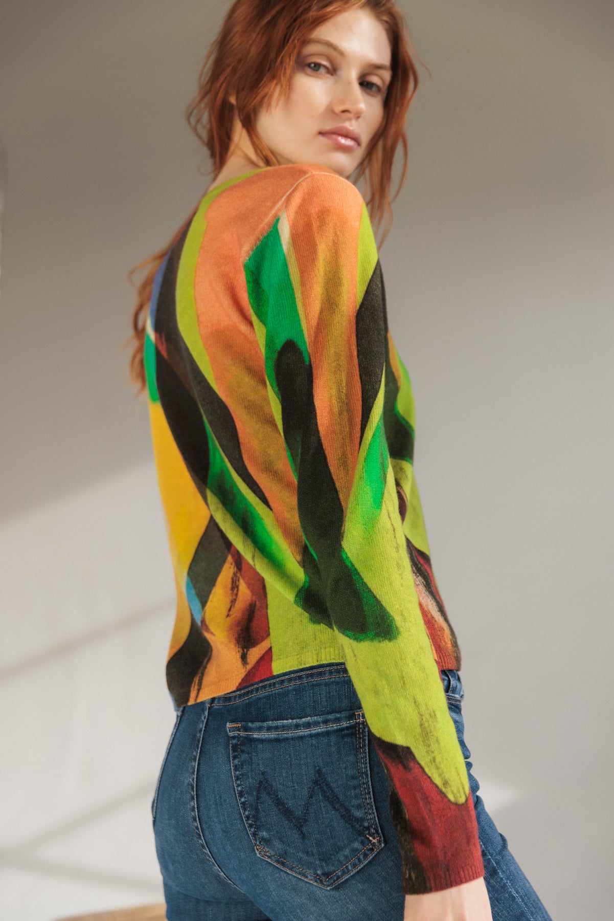 A woman is wearing a Catherine Gee Crewneck Cashmere Sweater - Stella Print design, featuring a vibrant, abstract pattern with green, red, yellow, blue, and black colors paired with blue jeans with visible stitching on the back pocket. The image is taken from the side, showing the back and part of the left arm of the sweater. 