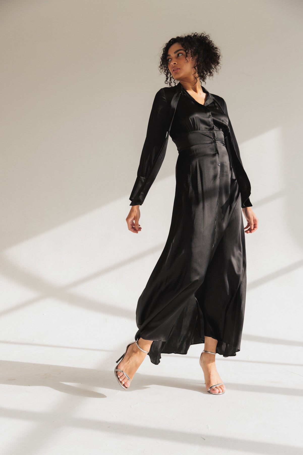 A woman wearing a black Catherine Gee Flou dress features a long, flowing silhouette made of a silky material that catches the light beautifully. It has long sleeves and a V-neckline, with a fitted waist that flares into a full skirt. The well-lit setting highlights the movement and elegance of the dress, creating a chic and sophisticated look.
