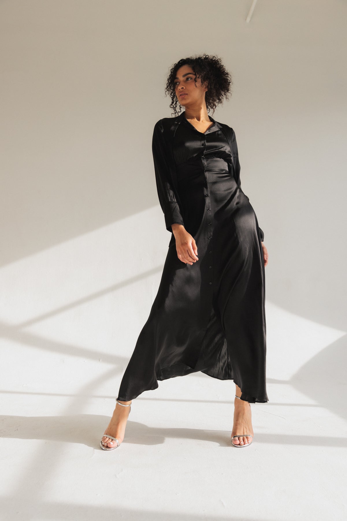 A woman wearing a black Catherine Gee Flou Dress. This elegant dress features a long, flowing silhouette made of a silky fabric that catches the light beautifully. It has a button-down front and long sleeves, adding to its sophisticated design. The minimalist background highlights the luxurious texture and fluid movement of the dress, making it a standout piece for any occasion.