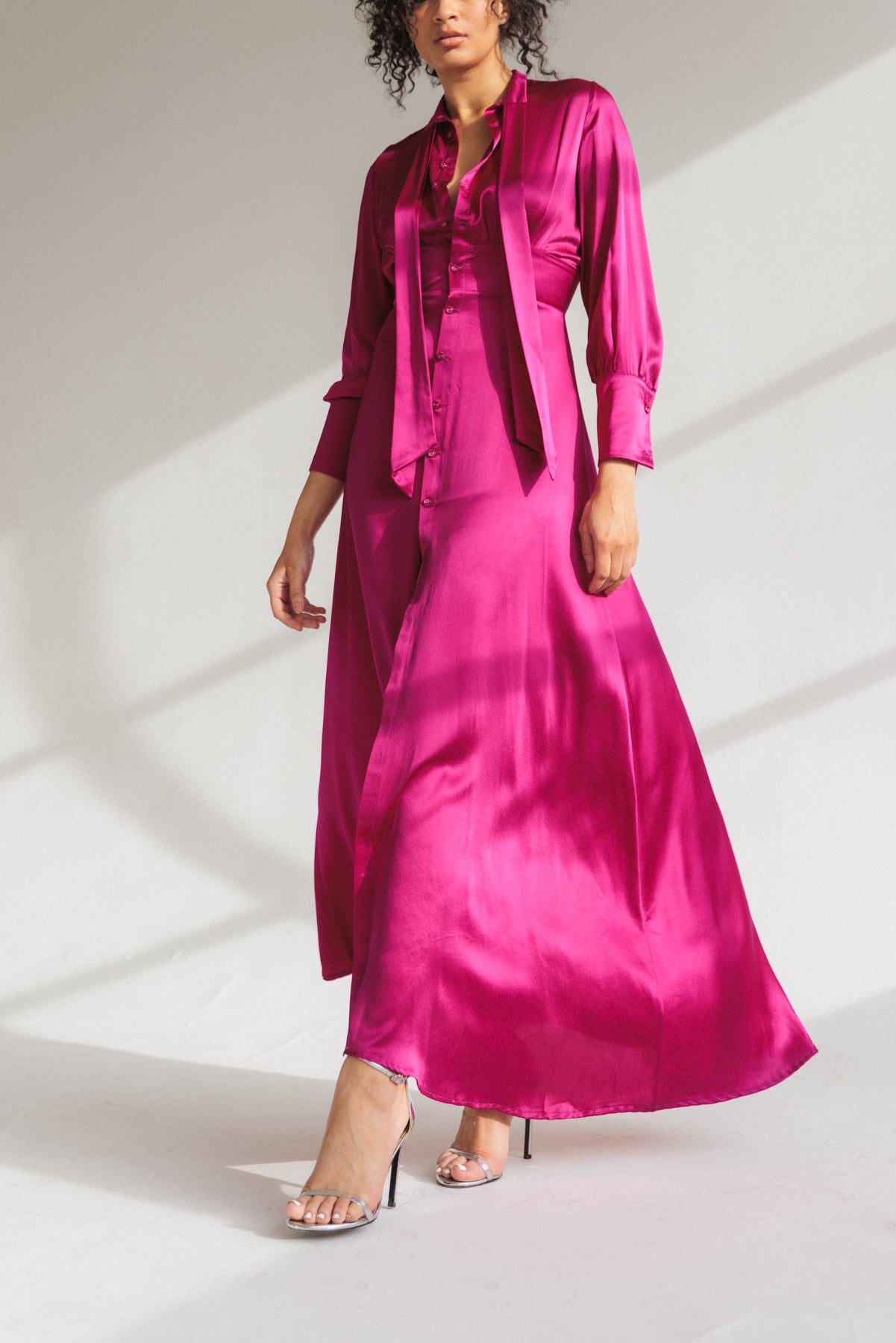 A woman wearing a Catherine Gee Electric Orchid Flou Dress. This vibrant dress features a silky, flowing fabric that reaches down to the ankles. It has long sleeves, a button-down front, and a tie detail around the neck paired with high-heeled, open-toe sandals. The rich color and luxurious fabric highlight the dress's elegance and sophistication, making it a standout piece.