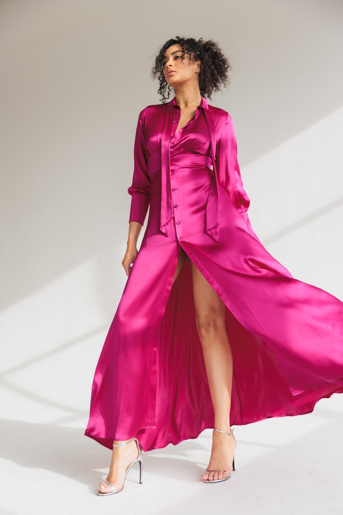 A woman wearing a Catherine Gee Electric Orchid Flou dress is a vibrant, bright pink color with a silky, satin-like texture. It features long sleeves, a button-down front, and a high slit that reveals one leg. The dress also has a tie detail at the neck, adding a sophisticated touch. She is standing in a well-lit space, with natural light casting soft shadows, highlighting the flow and sheen of the fabric.