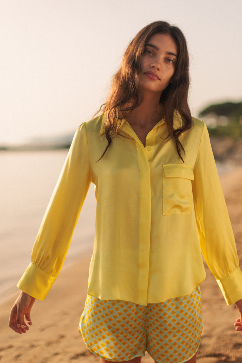 A woman standing on a beach wearing a Catherine Gee Jamie Yellow Blouse. The blouse is bright yellow with long sleeves and a single chest pocket on the left side. Made from a lightweight, slightly shiny fabric, it exudes a casual yet polished look. She pairs the blouse with yellow shorts featuring a blue polka dot pattern. The background includes a sandy beach and a blurred view of the sea, adding to the relaxed, summery vibe of the outfit.