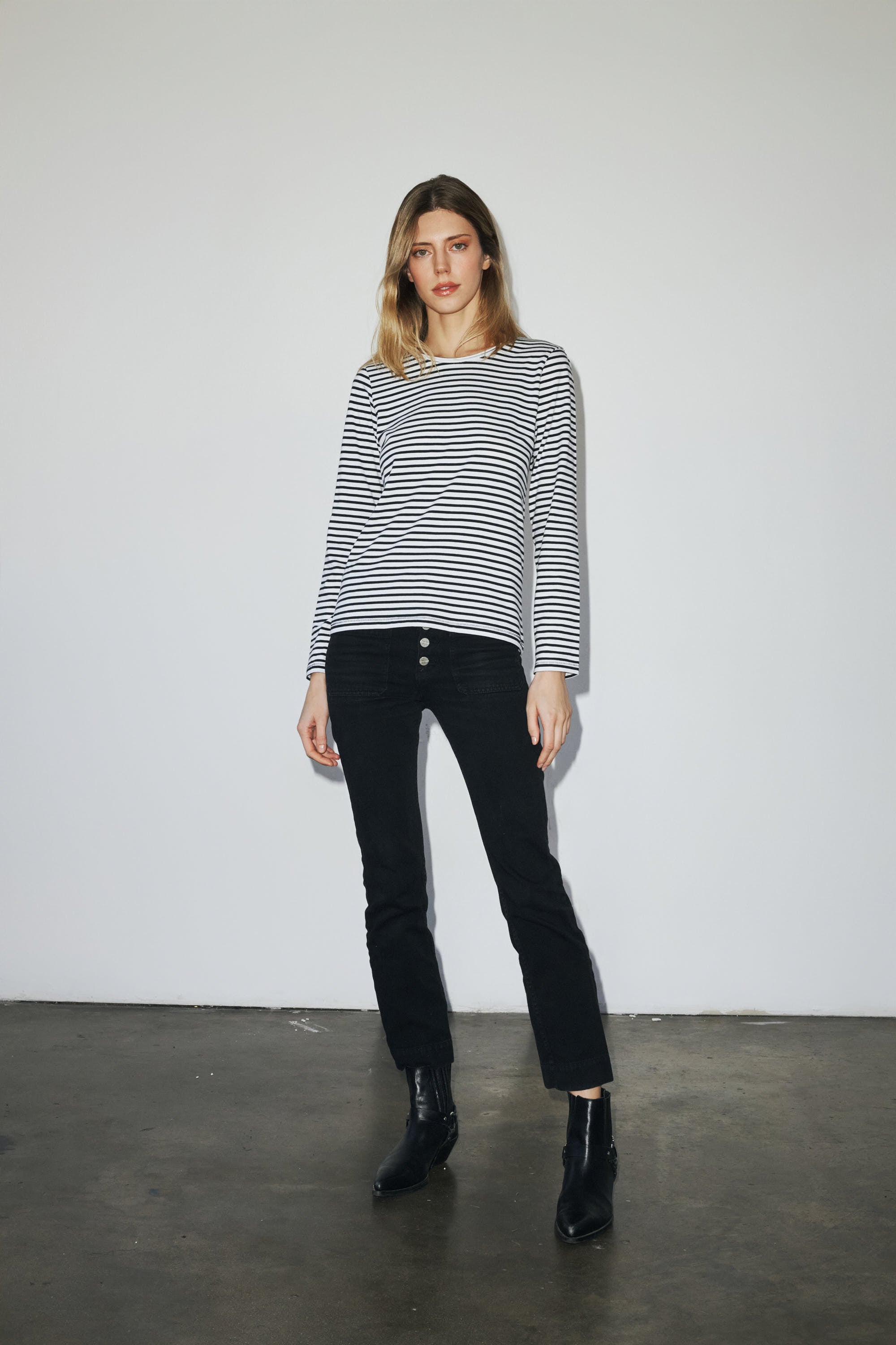 A woman is standing against a plain white background, wearing a black and white striped long-sleeve T-shirt and black pants paired with ankle boots. The outfit is casual and stylish, suitable for everyday wear showcasing the Catherine Gee black and white striped long-sleeve T-shirt, highlighting its fit and style.