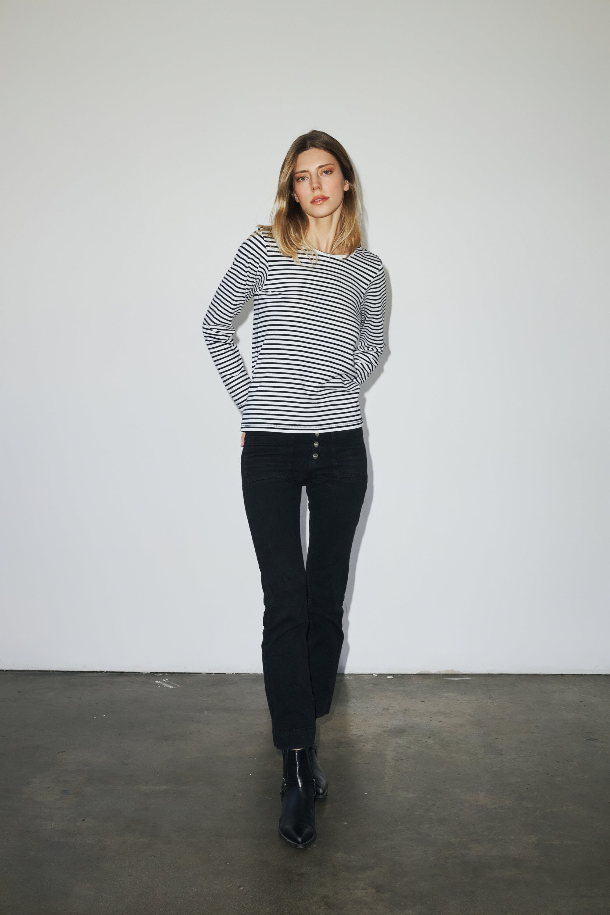 A woman is standing against a plain white background, wearing a black and white striped long-sleeve T-shirt by Catherine Gee. She is also wearing black pants and black ankle boots. The simplicity of the background focuses attention on the T-shirt's classic design and fit, making it the standout piece in the outfit.