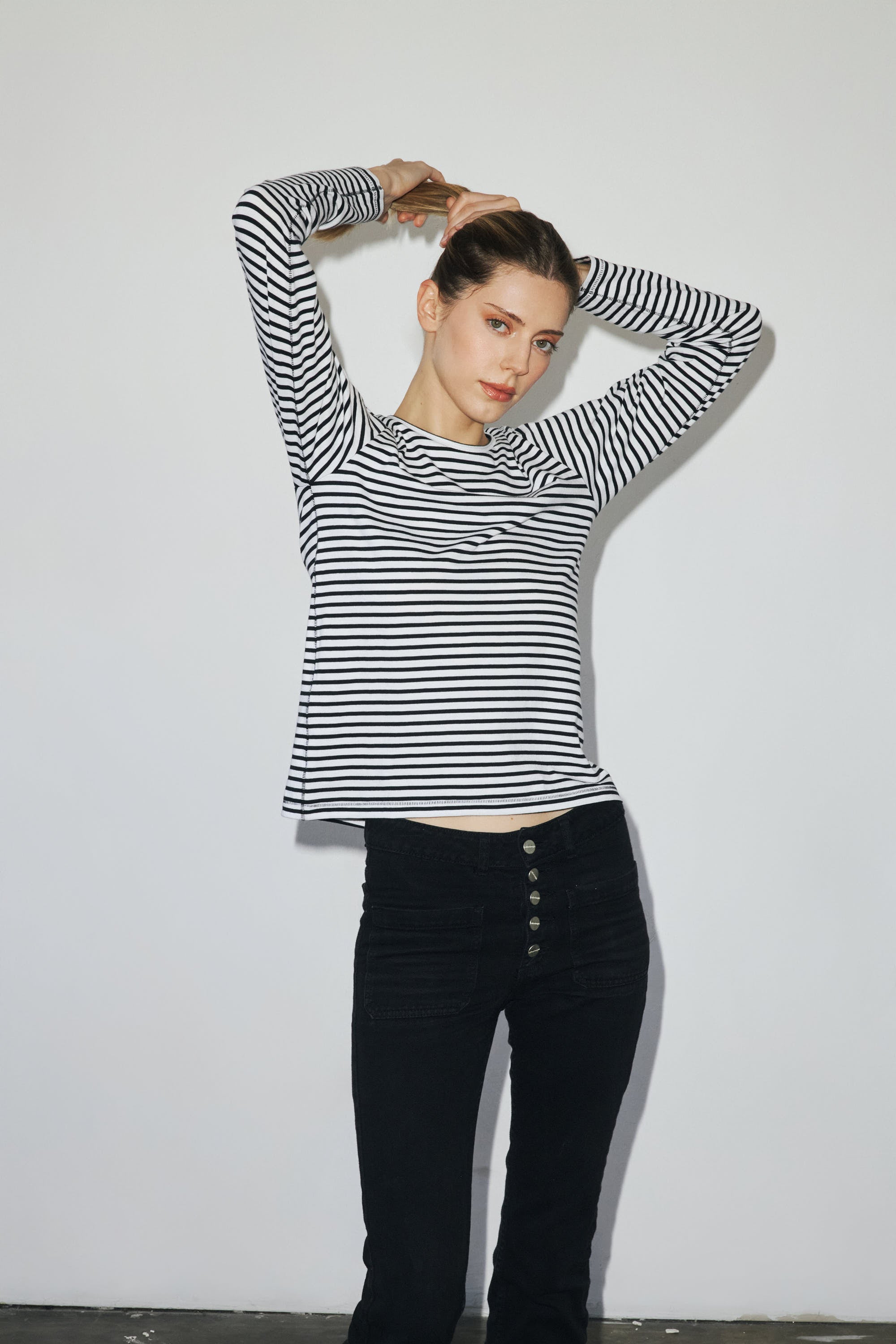 A woman is wearing a black and white striped long-sleeve T-shirt from Catherine Gee. She is posing with arms raised, and hands behind the head, highlighting the fit and design of the T-shirt. The background is plain white, drawing focus to the outfit's style and details.