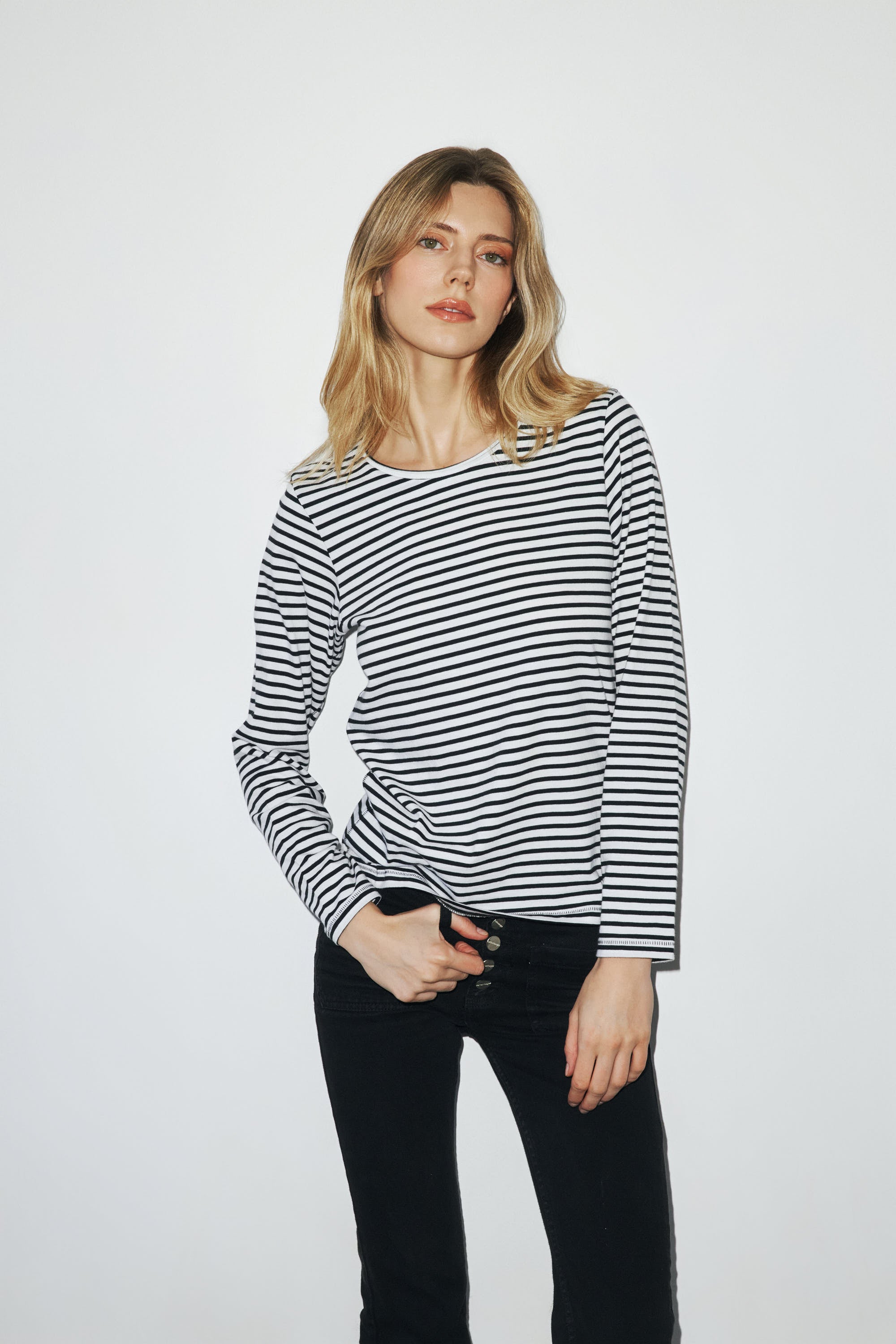 A woman is wearing a black and white striped long-sleeve T-shirt by Catherine Ge features horizontal stripes and a relaxed fit. She is also wearing black pants and has one hand resting on the waistband of the pants in a plain and light-colored background.