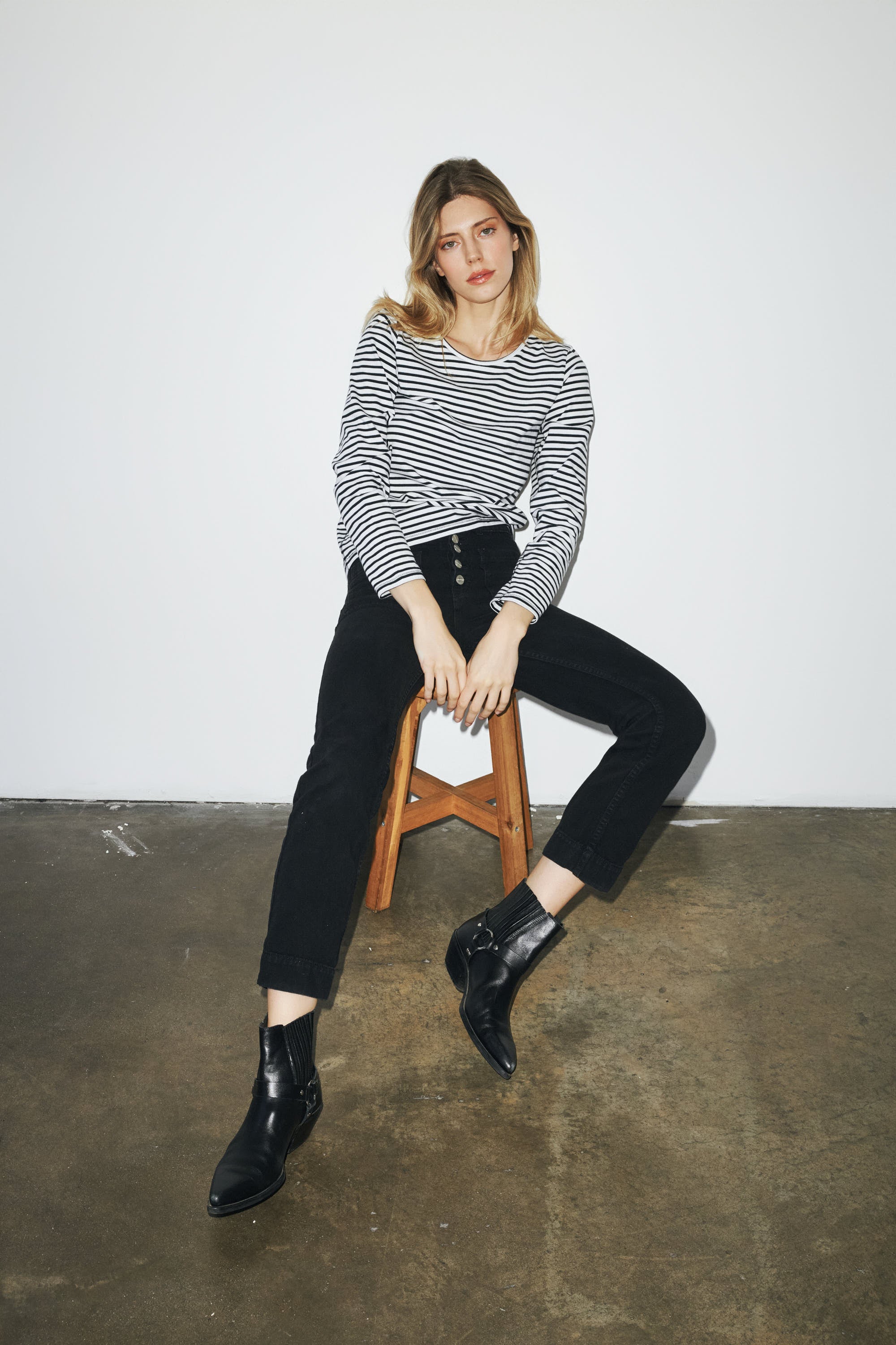 A woman is sitting on a wooden stool against a plain white wall, wearing a black and white striped long-sleeve T-shirt by Catherine Gee. She is also dressed in black high-waisted pants and black ankle boots. The setting appears to be a minimalist indoor space with a concrete floor.