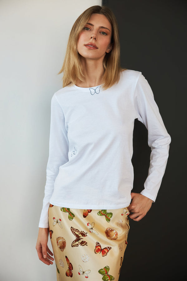 A woman wearing a Catherine Gee Embroidered Cotton Long Sleeve Butterfly white t-shirt features delicate butterfly embroidery near the neckline and the lower left side paired with a skirt with a colorful butterfly print, which complements the t-shirt's minimalist design. The overall look is elegant and charming, perfect for those who appreciate subtle, artistic details in their clothing.