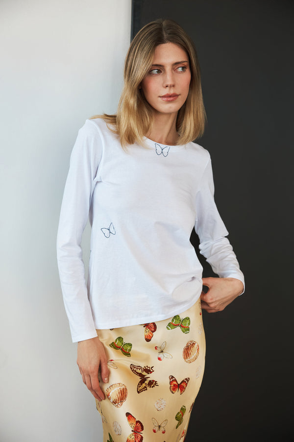 A woman wearing a Catherine Gee Embroidered Cotton Long Sleeve Butterfly white t-shirt with small, delicate butterfly embroidery on the chest area paired with a colorful butterfly print skirt, which complements the butterfly theme of the t-shirt. The background is a simple, two-tone wall, which helps to highlight the clothing, and showcases a coordinated outfit with a nature-inspired theme, emphasizing the complex embroidery and print details.