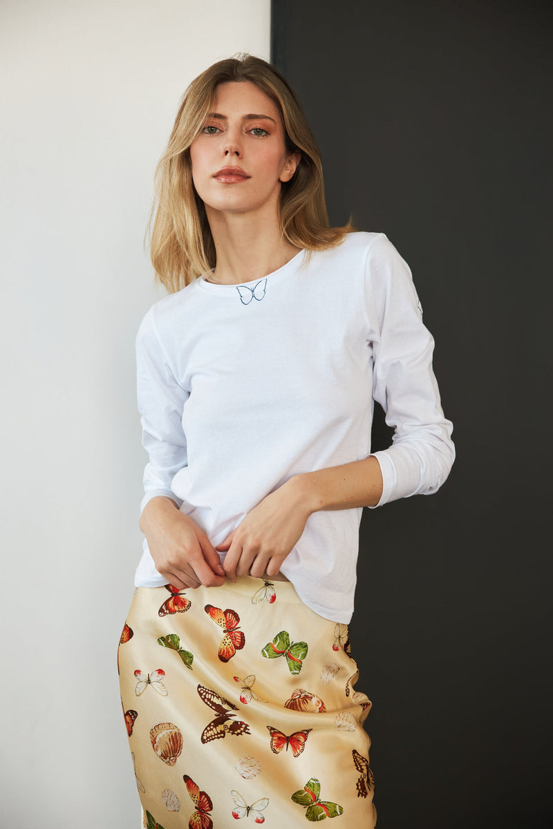 A woman wearing a Catherine Gee Embroidered Cotton Long Sleeve Butterfly white t-shirt features a small, delicate butterfly embroidery near the neckline. She also wears a beige skirt decorated with colorful butterfly prints, creating a cohesive and visually appealing outfit. The combination of embroidered details on the t-shirt and the vibrant prints on the skirt highlights the elegance and charm of the shirt.