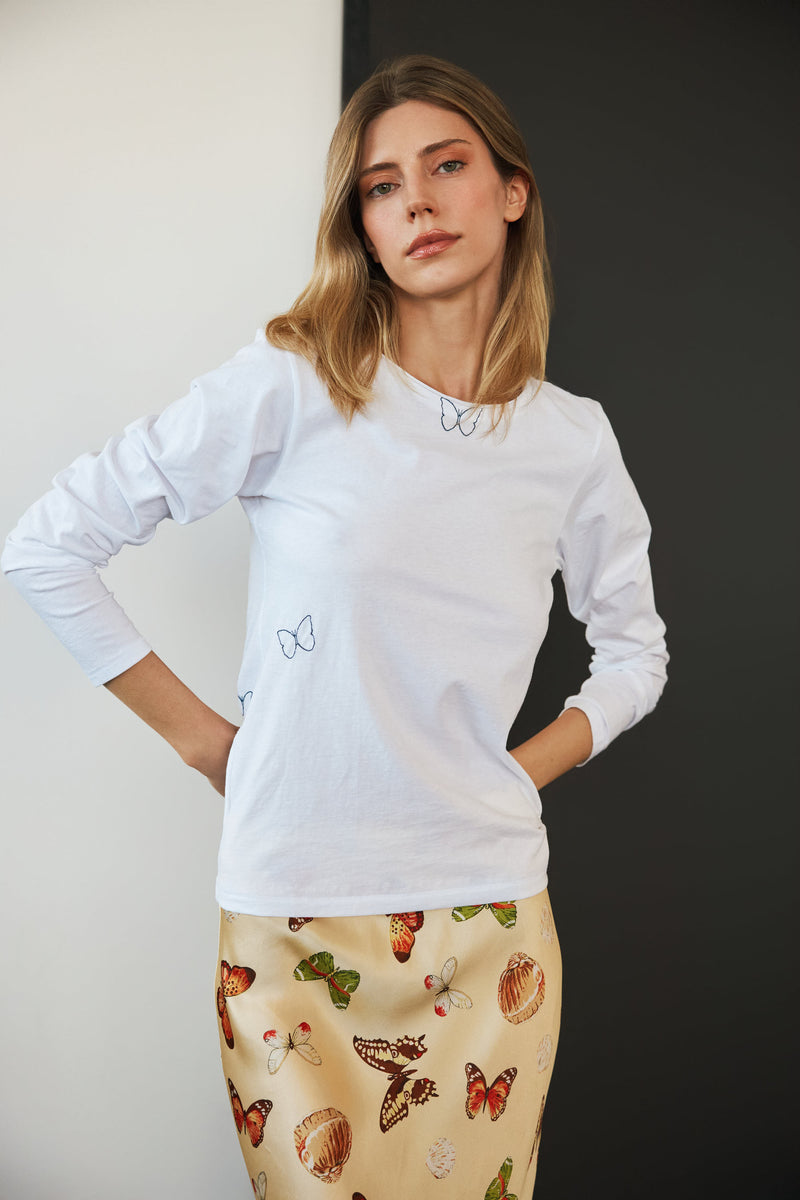 A woman wearing a Catherine Gee Embroidered Cotton Long Sleeve Butterfly T-Shirt. The shirt is white with simple butterfly embroidery on the chest and near the neckline paired with a colorful butterfly print skirt highlighting the delicate and nature-inspired design of the clothing, making it interesting for those who appreciate detailed embroidery and fashion.