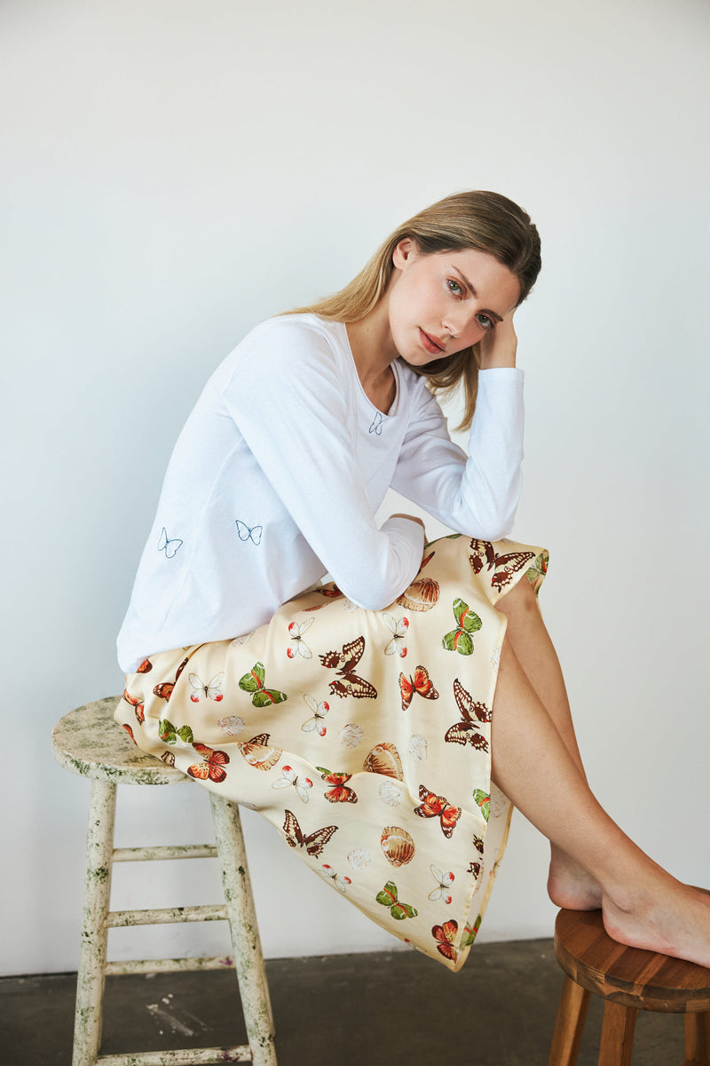 A woman seated on a stool, wearing a Catherine Gee Embroidered Cotton Long Sleeve Butterfly white t-shirt features delicate butterfly embroidery on the sleeves and pairs with a cream-colored skirt decorated with a colorful butterfly pattern, creating a cohesive, nature-inspired outfit. The minimalist background highlights the detailed embroidery and prints, making the ensemble both charming and elegant.