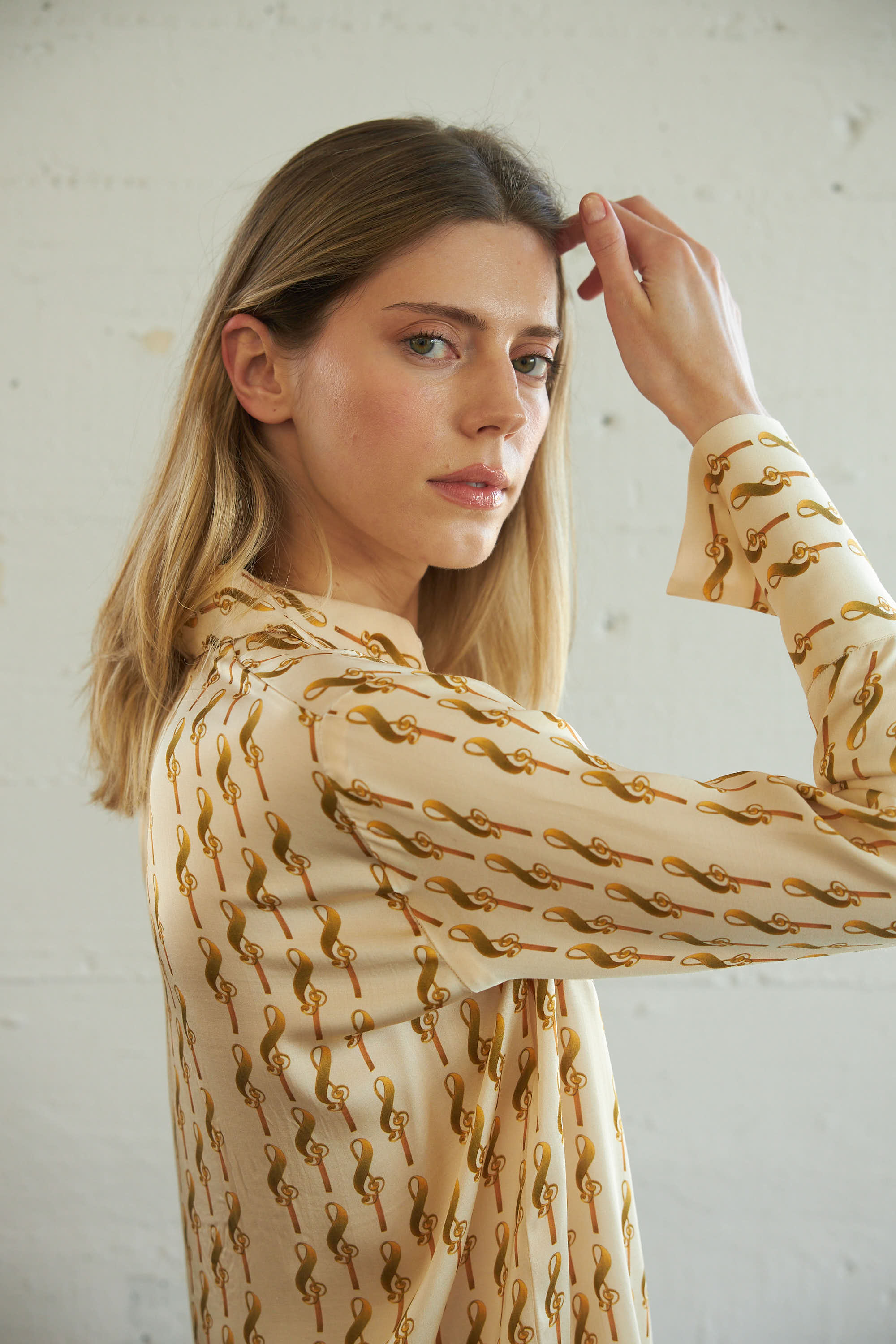 A woman is wearing a Catherine Gee Dakota Blouse in a cream color with a gold treble clef pattern. The blouse features a relaxed fit with long sleeves and is paired with blue jeans. She has shoulder-length blonde hair and is touching her head with one hand, showcasing the sleeve and pattern.