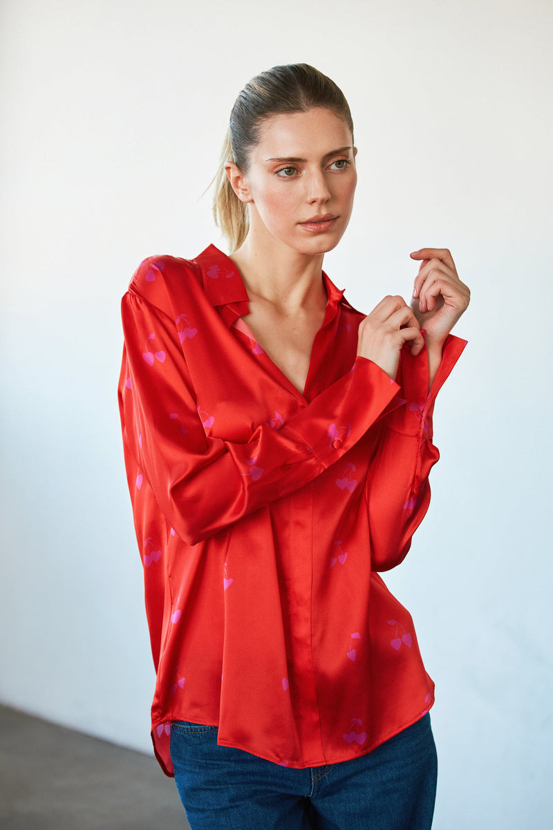 A woman wearing a Catherine Gee Daria French Cuff Silk-Cherry Heart Blouse features a vibrant red silk blouse with a subtle heart pattern, and a French cuff design, and has a relaxed, slightly oversized fit. She is also wearing blue jeans, which highlight the luxurious texture and elegant design of the blouse, making it interesting for fashion enthusiasts.