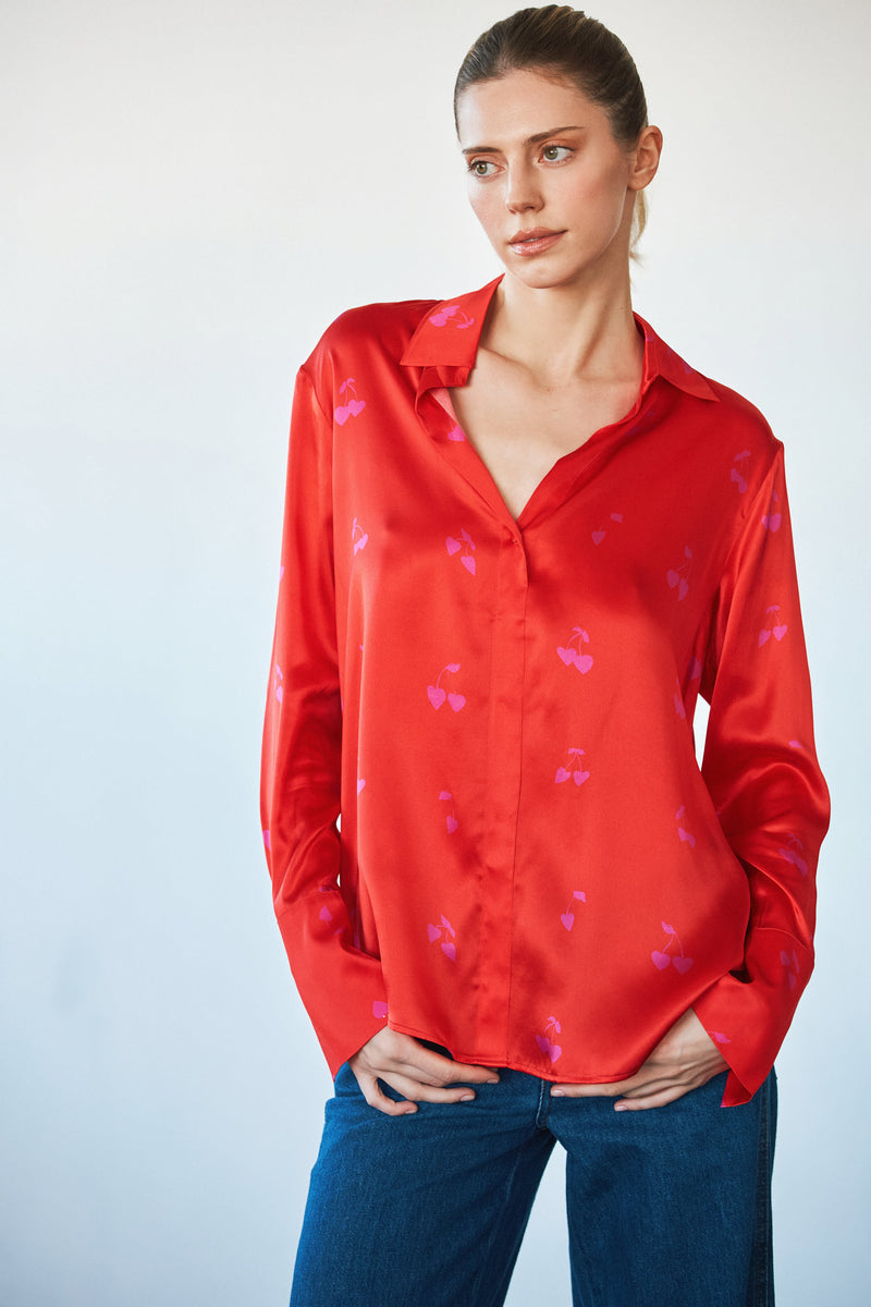 Woman wearing a Catherine Gee Daria French Cuff Silk Blouse in Cherry Heart. The blouse is a vibrant red color with a subtle pattern of small pink hearts. It has a classic collar and long sleeves with French cuffs paired with blue jeans. The blouse appears to be made of a smooth, shiny silk material, giving it a luxurious look.