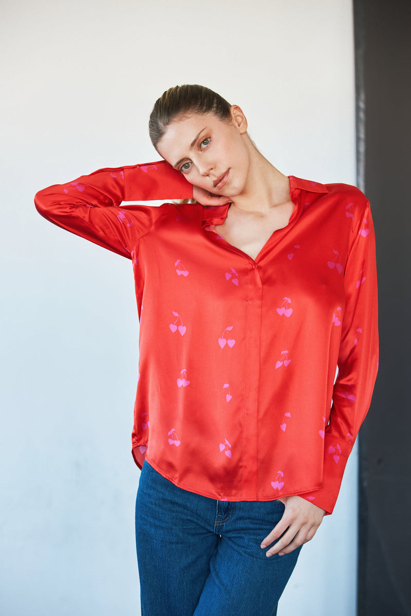 A woman wearing a Catherine Gee Daria French Cuff Silk-Cherry Heart Blouse features a vibrant red color with a cherry heart pattern, V-neckline, and long sleeves with French cuffs. She poses with one hand behind the head and the other hand resting on the hip on a simple neutral background which helps to highlight and showcase the blouse.