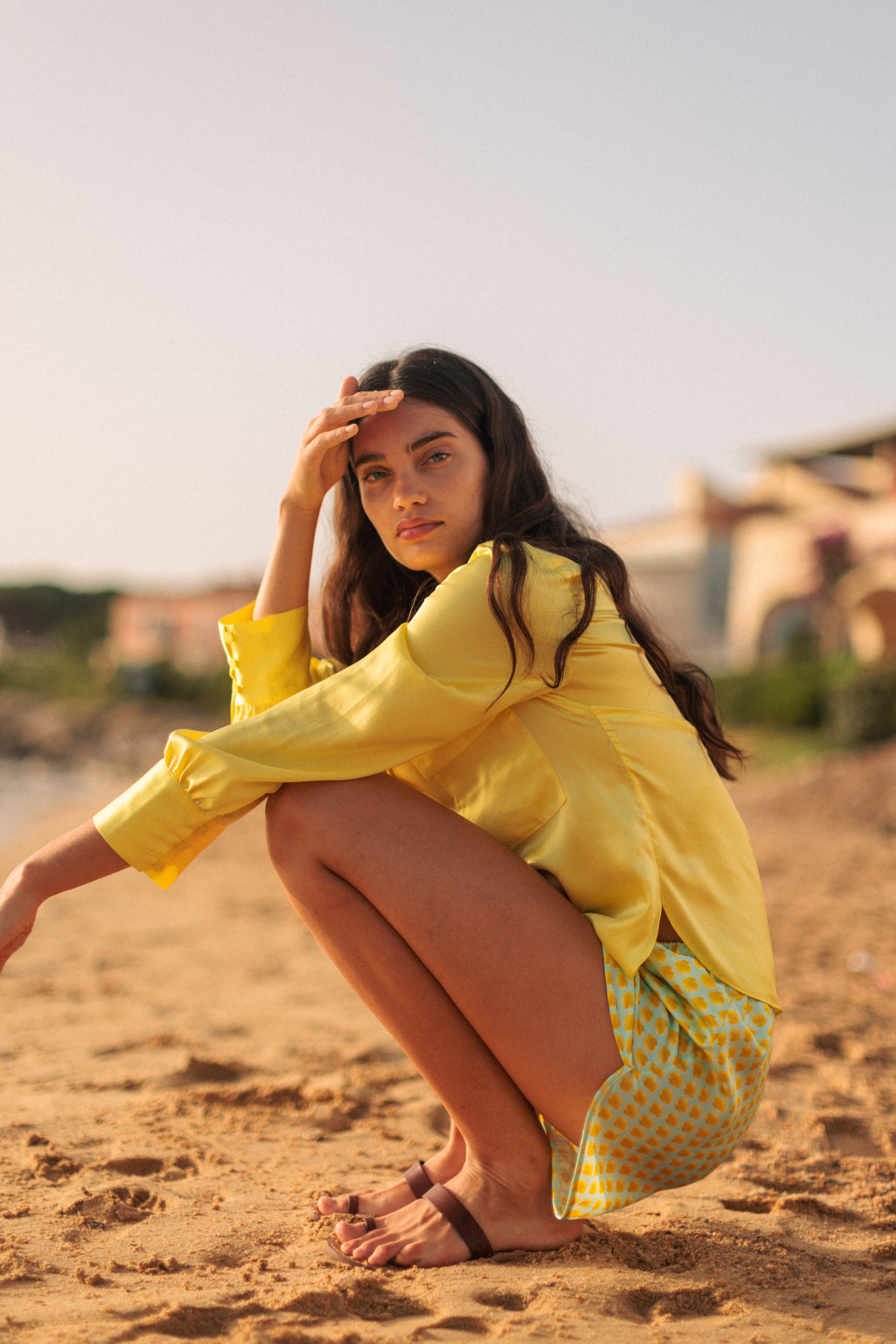 A woman with long, wavy hair is squatting on a sandy beach, wearing a Catherine Gee Jamie Yellow Blouse and sandals. The blouse is long-sleeved and loose-fitting, featuring a bright, sunny hue that complements the beach setting. The background includes blurred buildings and greenery, enhancing the relaxed, summery atmosphere of the scene.