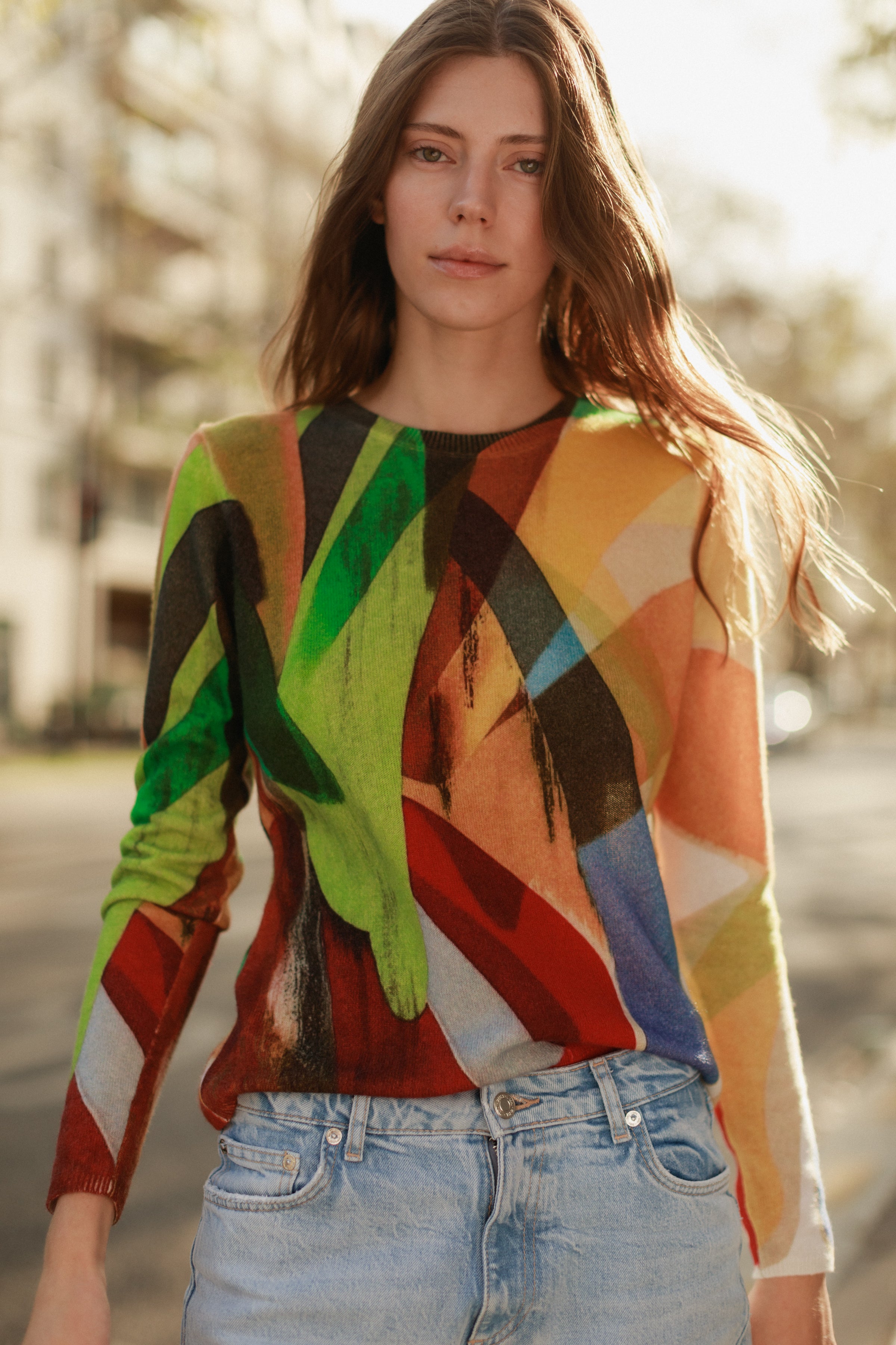 A woman is wearing a Catherine Gee Crewneck Cashmere Sweater - Stella Print design features a vibrant, abstract pattern with various colors including green, red, yellow, blue, and black paired with light blue jeans. The background is an outdoor urban setting with blurred buildings and trees, suggesting a casual, stylish look.