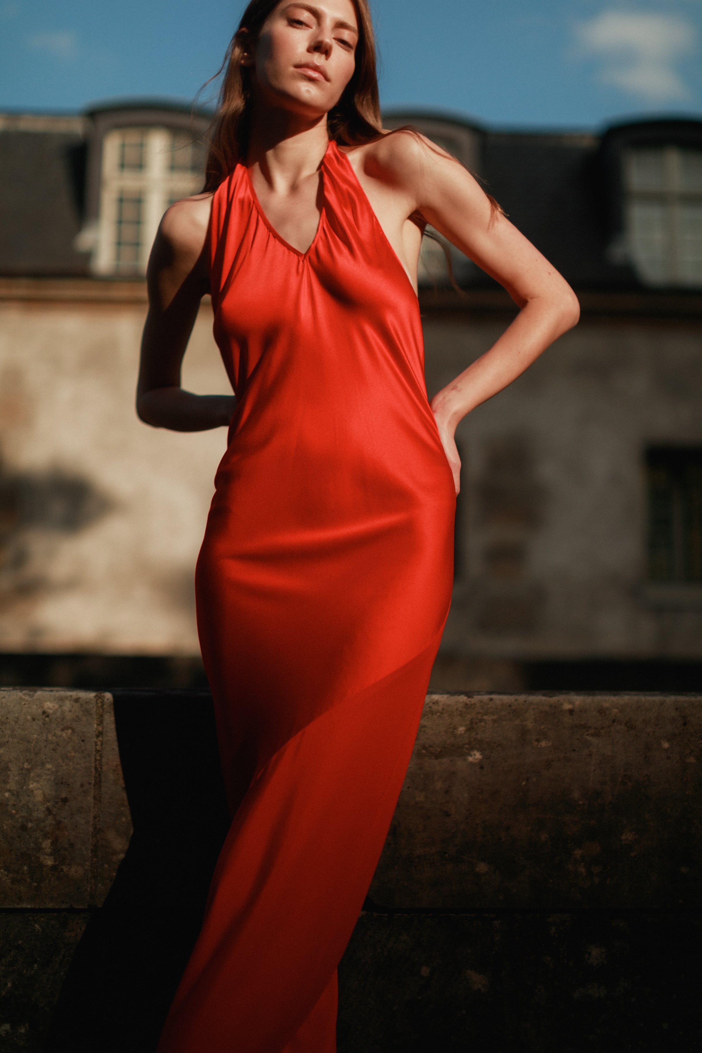A woman wearing a Catherine Gee Diane High-Risk Red striking dress features a vibrant red color and a halter neckline. The smooth, slightly shiny fabric drapes elegantly down to the ankles, highlighting the fitted silhouette with a blurred building in the background emphasizing the boldness and sophistication of the dress.