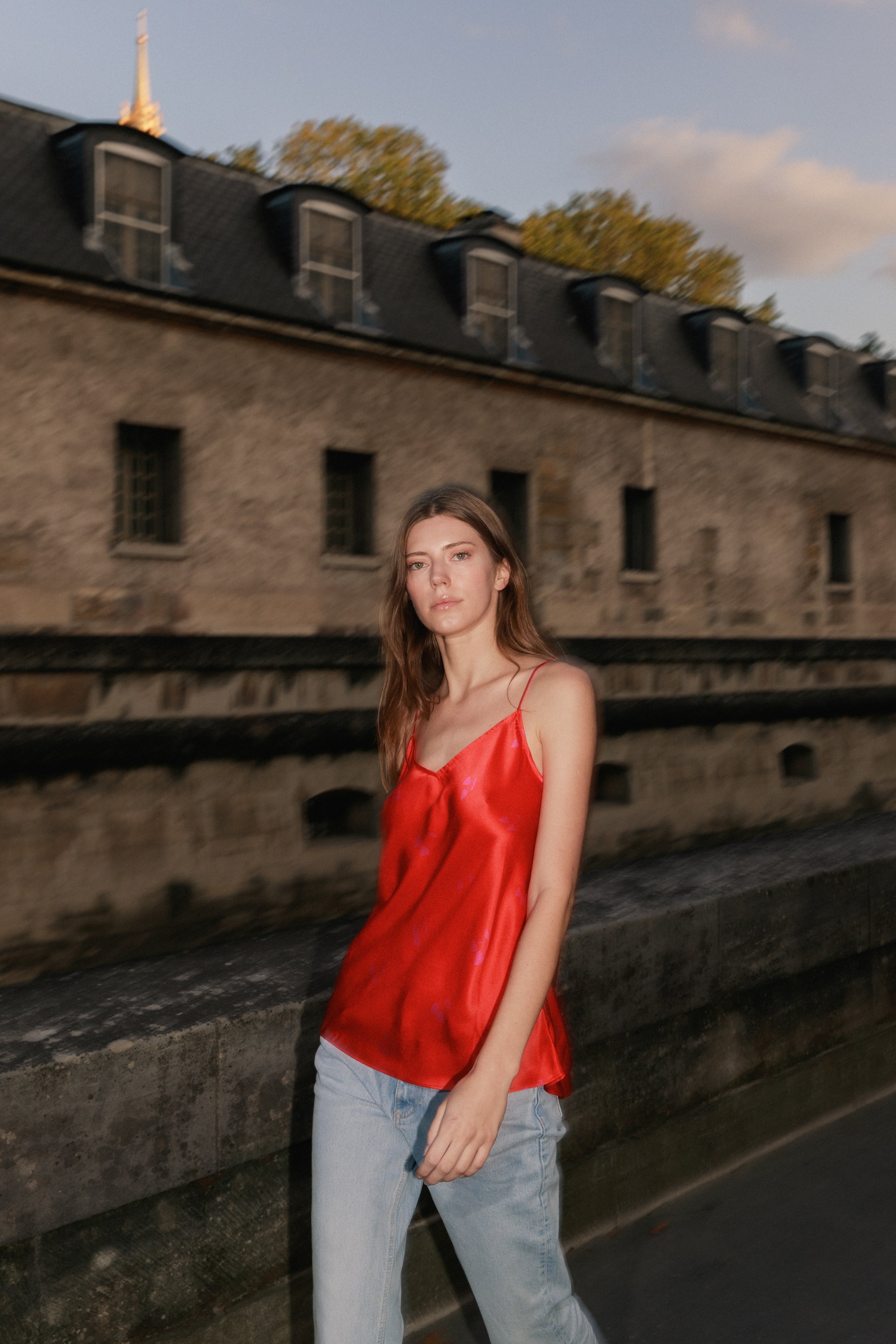 A woman is standing outdoors in front of a historic stone building in a clear sky with some clouds and trees, wearing a red Catherine Gee Basic Cami - Cherry Heart and light blue jeans.