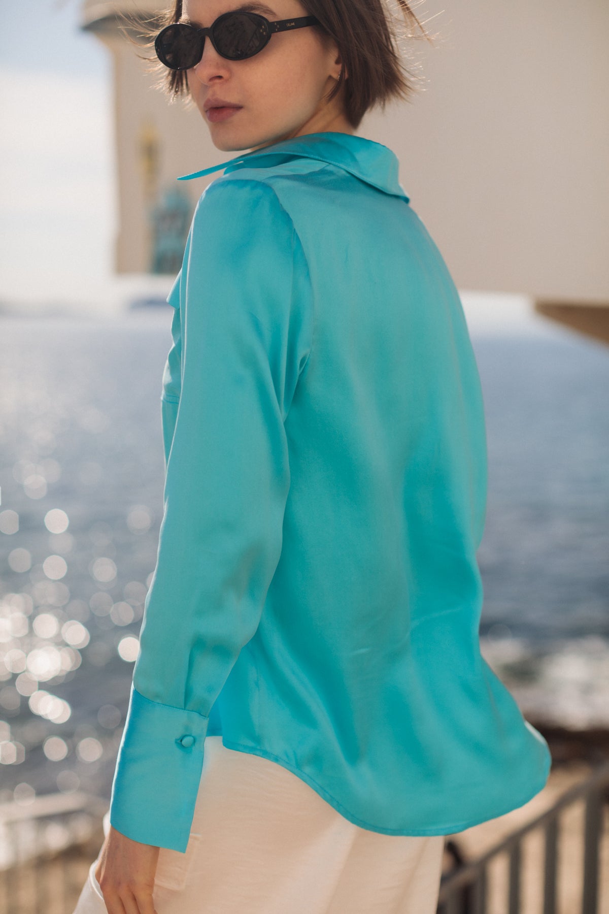 A woman wearing a Catherine Gee Daria French Cuff Silk Blouse Fiji Blue. The blouse has a smooth, shiny texture and features a classic collar and French cuffs with buttons. She also stands outdoors near a body of water, with sunlight reflecting off the water's surface, creating a sparkling effect, emphasizing the blouse's vibrant color and elegant design.
